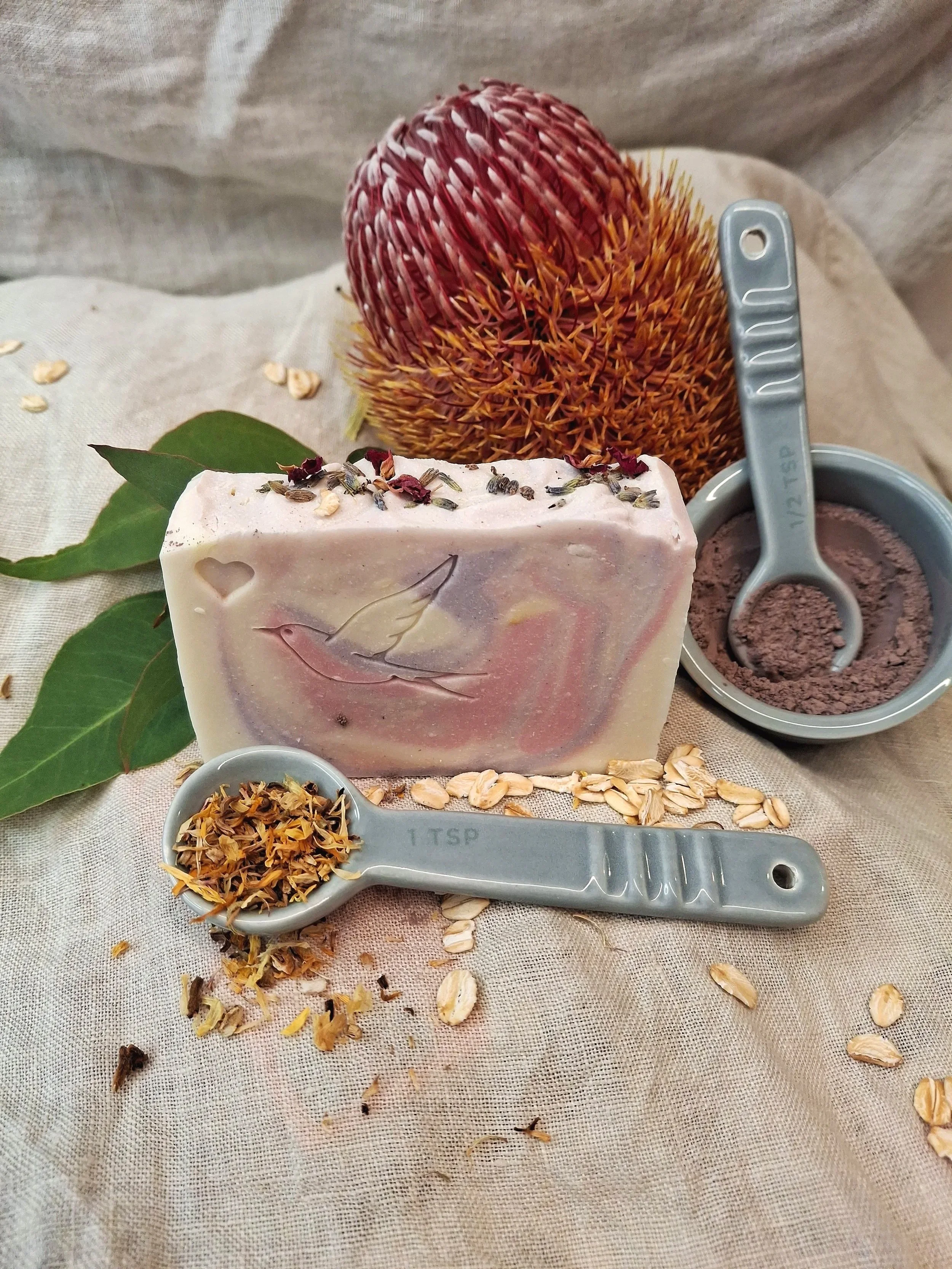 A bar of soap with dried flower petals on top, a metal scoop filled with brown powder, a large dried flower, and two small dishes with dried herbs and oats, all arranged on a beige fabric surface. Tallow soap perth western australia.