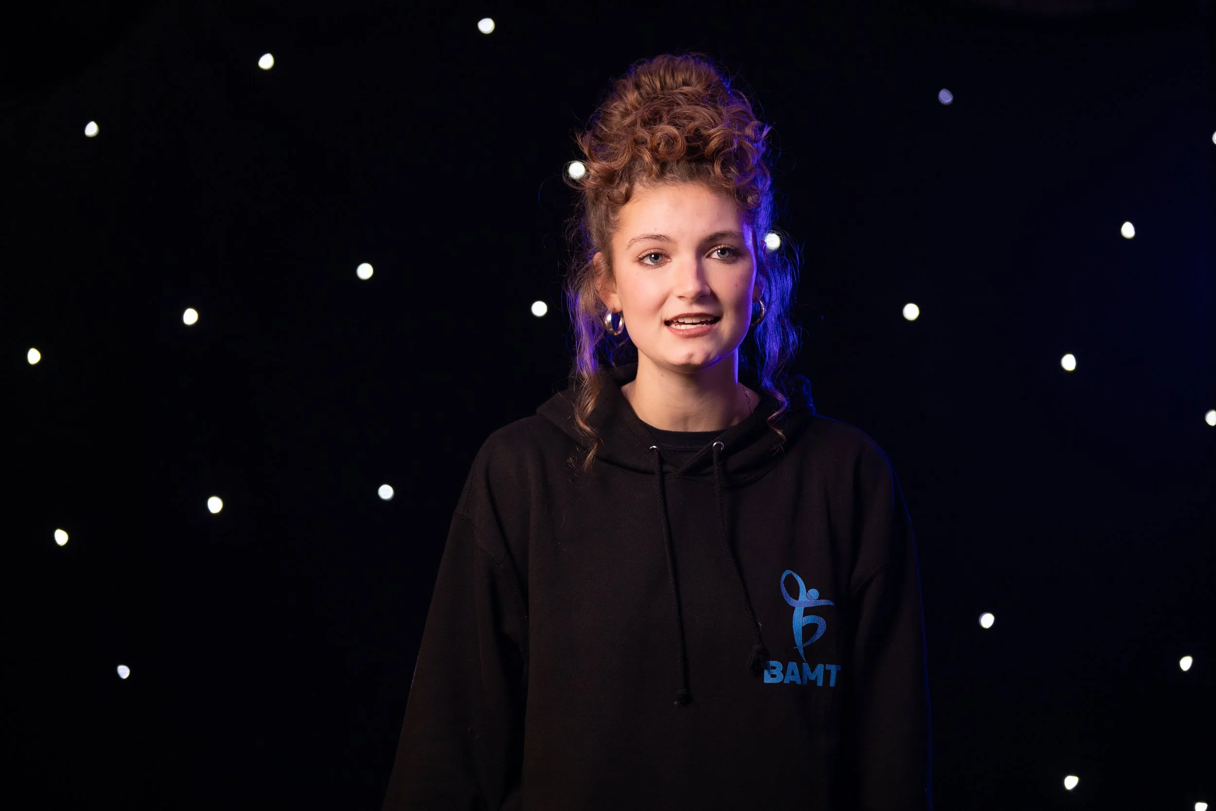 A young female BAMT student with curly hair styled in an updo, wearing hoop earrings and a black hoodie with blue text, standing against a dark background with small white lights.