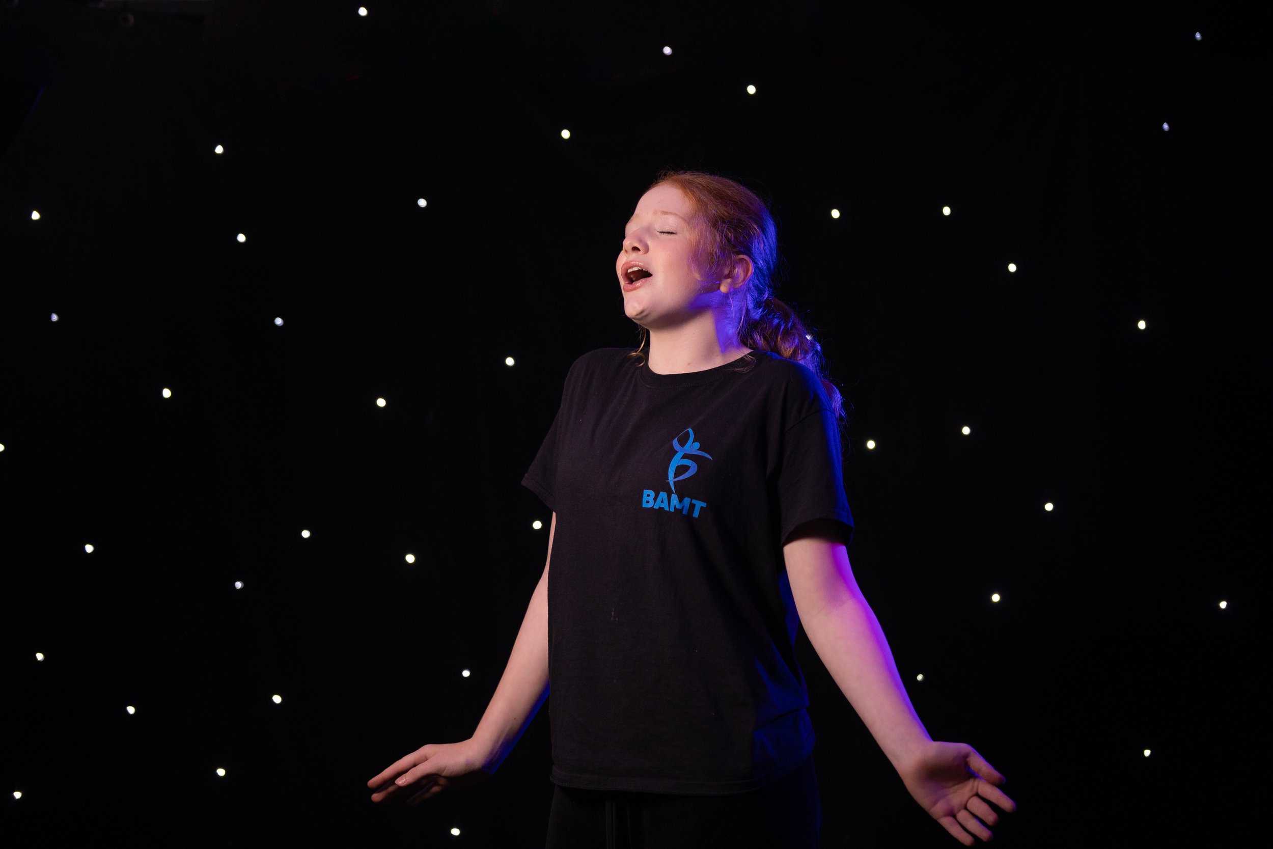 A young female BAMT student with red hair singing or performing on stage, standing with eyes closed and arms relaxed, against a black backdrop with small white lights.