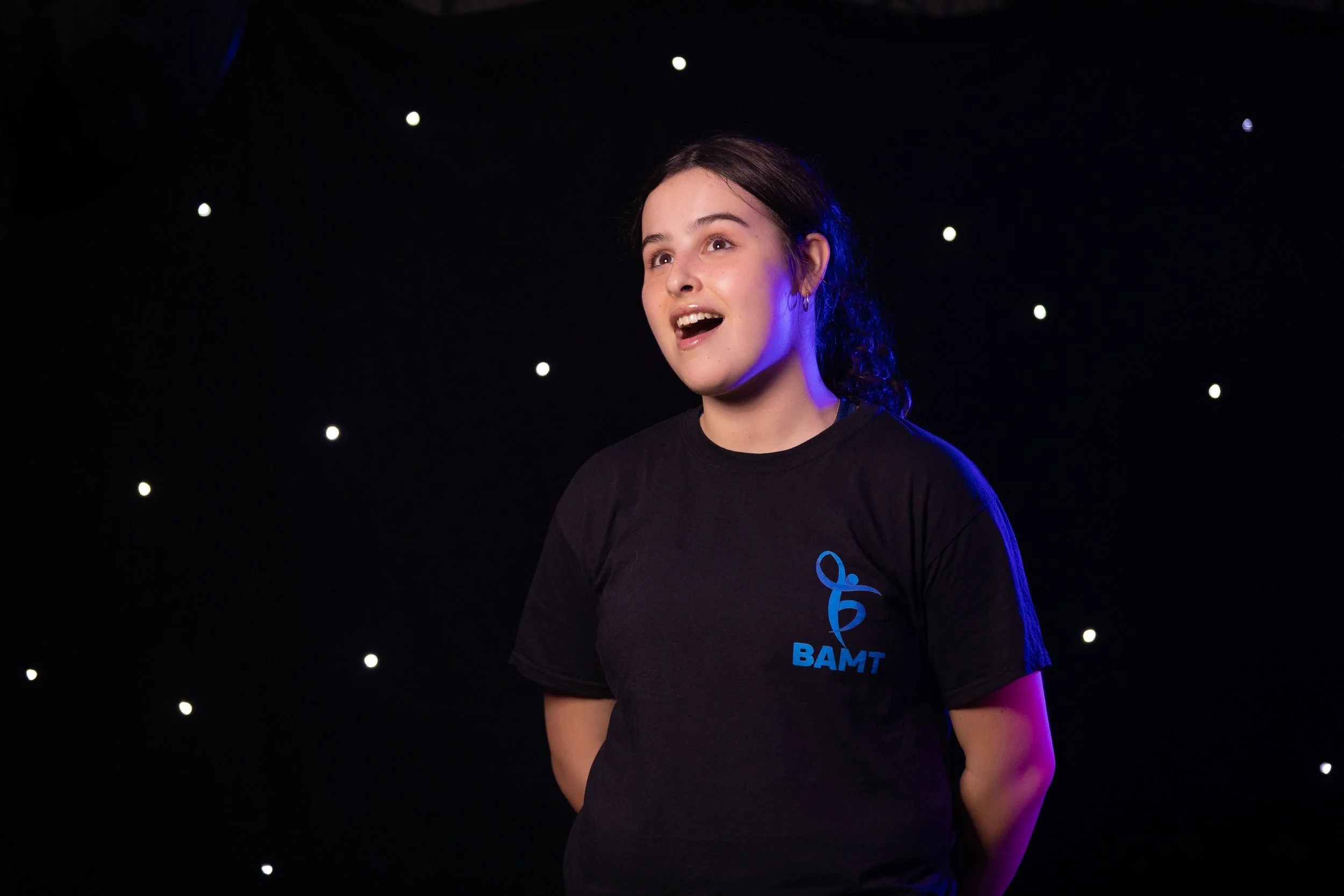 A young female BAMT student with dark hair pulled back, wearing a black T-shirt with a blue logo and text, standing against a black background with small white lights.