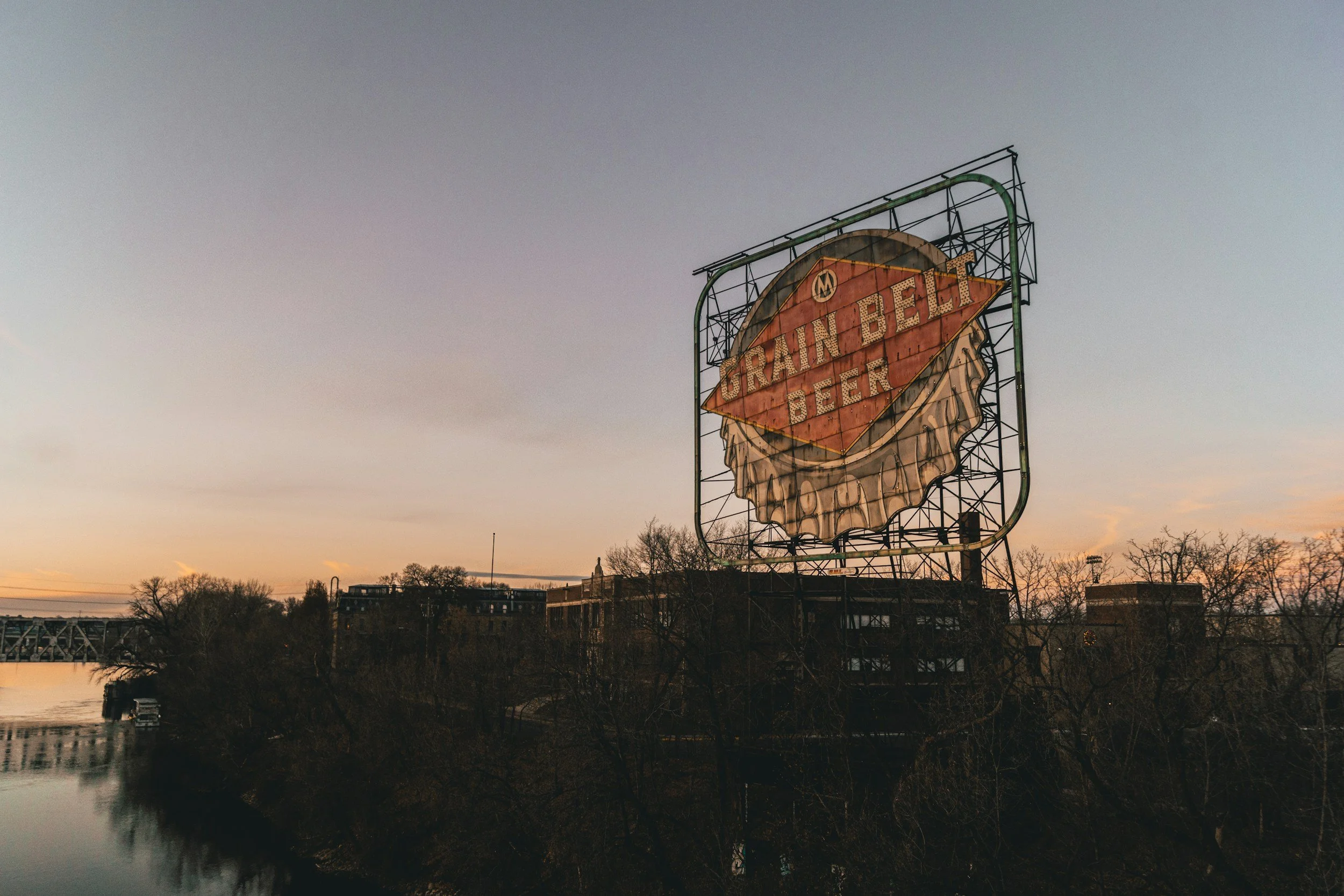 An old neon sign for Rain Belt Beer is seen against the evening sky, with trees and a river below.