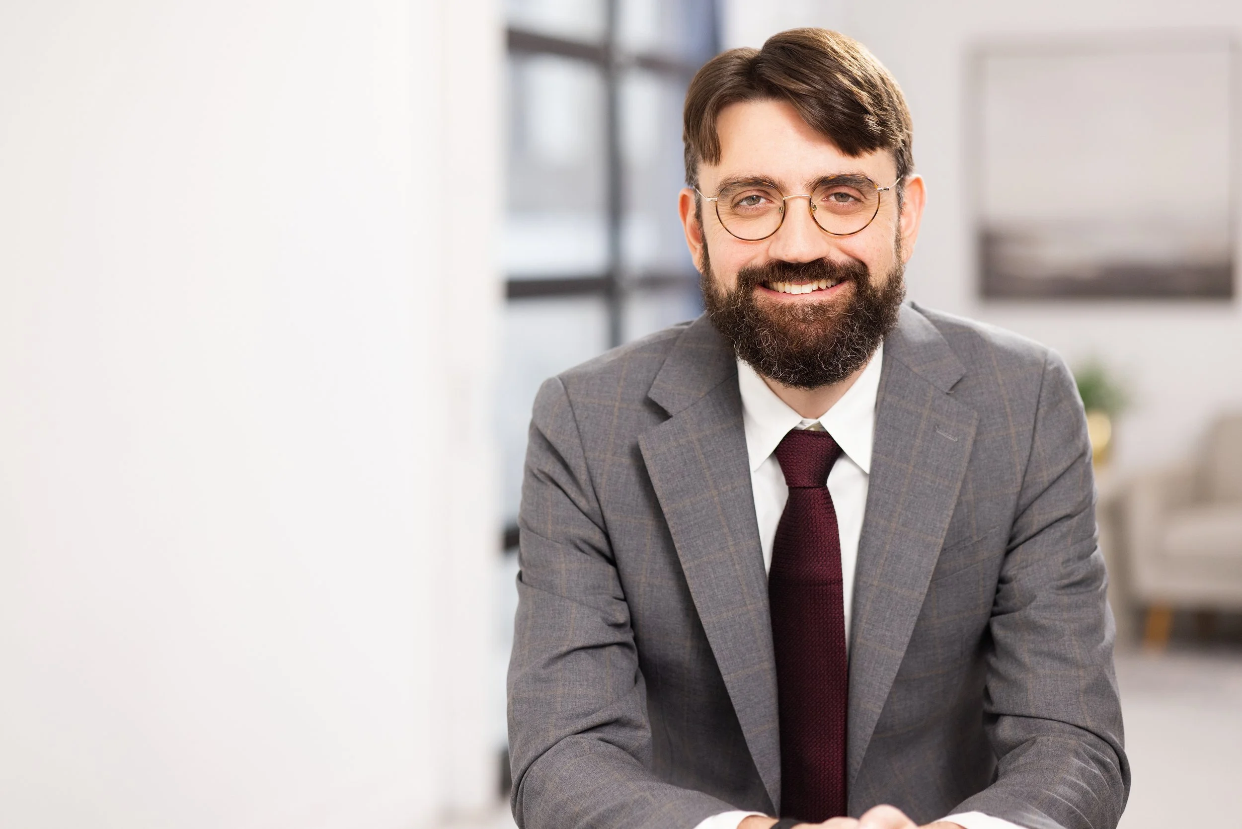 Man with glasses and a beard smiling in an office setting, wearing a gray suit and maroon tie.