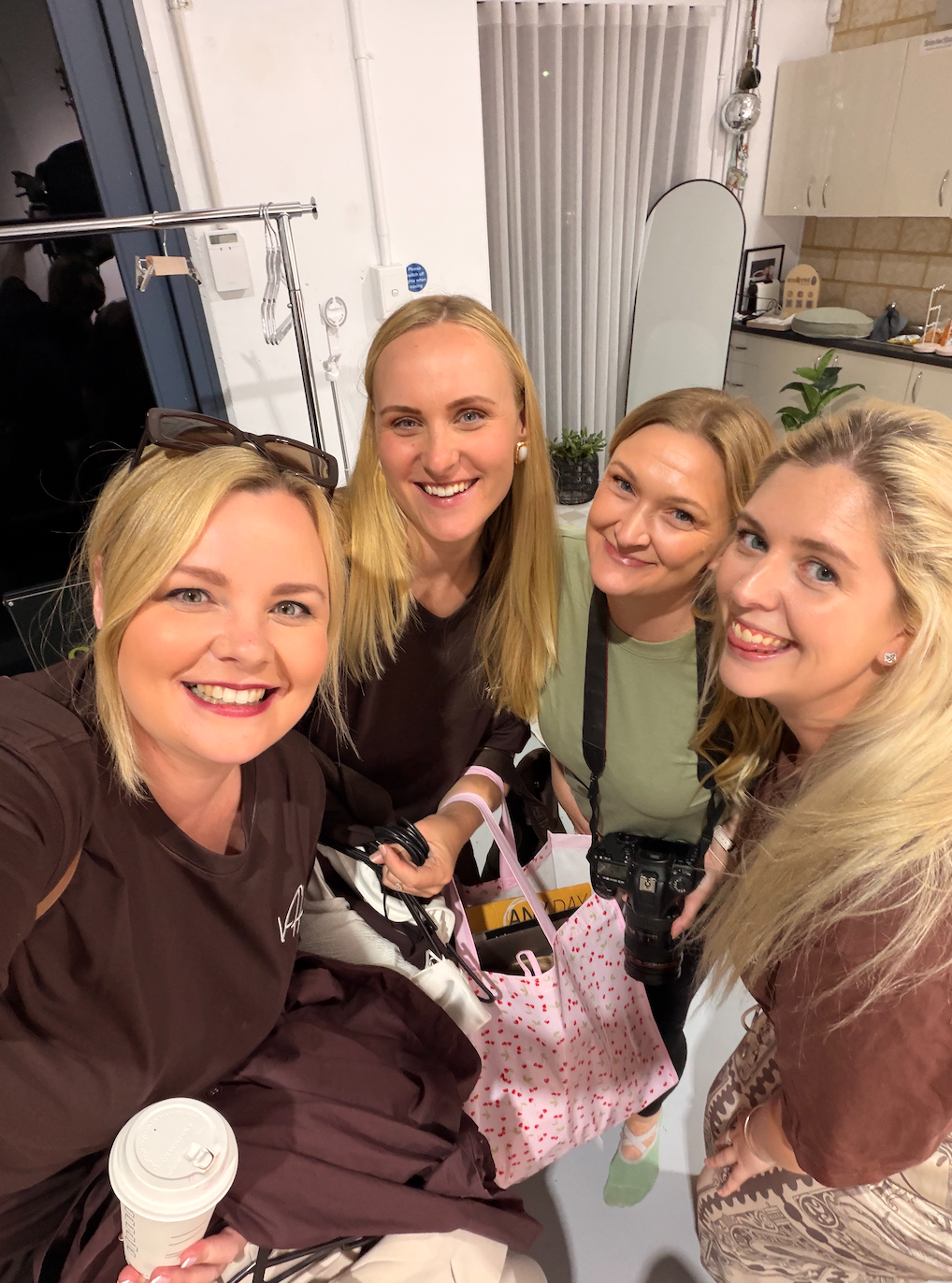 Group of four women smiling and taking a selfie in an indoor setting, with one woman holding a coffee cup and another holding a camera.