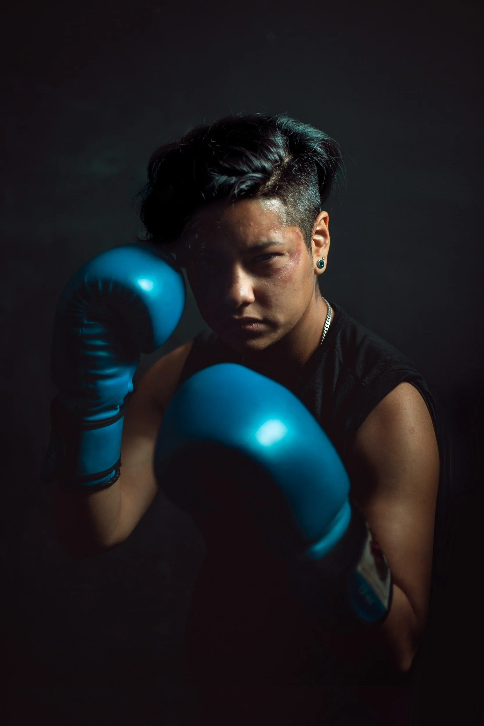 A person with short dark hair, wearing blue boxing gloves, a black sleeveless shirt, and earrings, is posed with one glove near the face and the other extended forward, against a dark background.