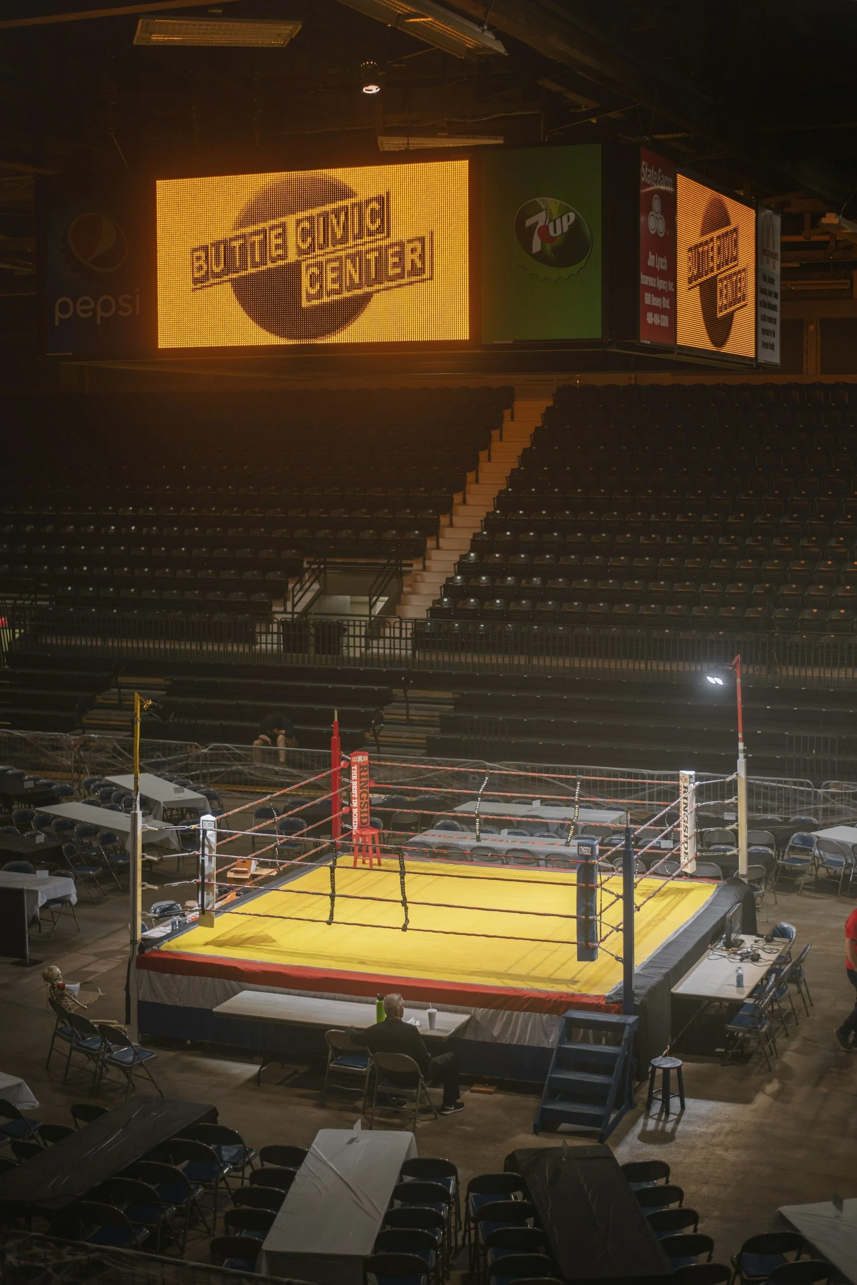 Indoor sports arena with a boxing ring in the foreground, surrounded by seating and tables. Large digital screens behind the boxing ring display advertisements for Butt Cave, 7Up, and Pepsi, with the arena dimly lit.
