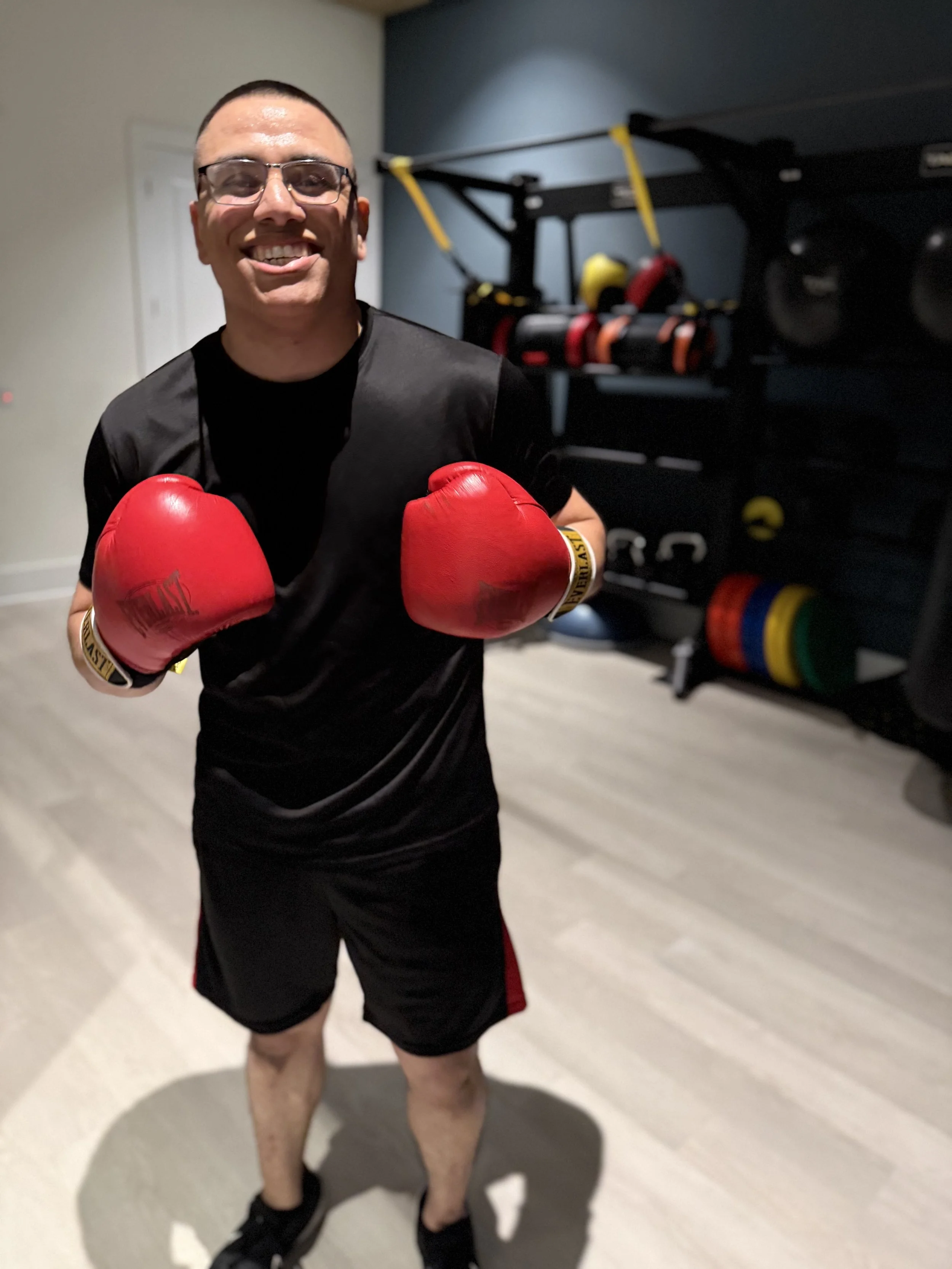 A young man with glasses and short hair wearing a black T-shirt and shorts, standing in a room, smiling, and wearing red boxing gloves. Behind him, fitness equipment including a rack with colorful weight plates and resistance bands.