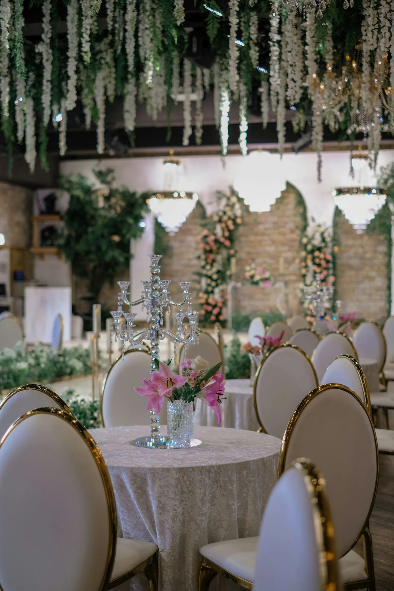 Wedding reception setup with round tables, floral centerpieces, and hanging floral decorations, chandeliers, and elegant chairs with gold trim.