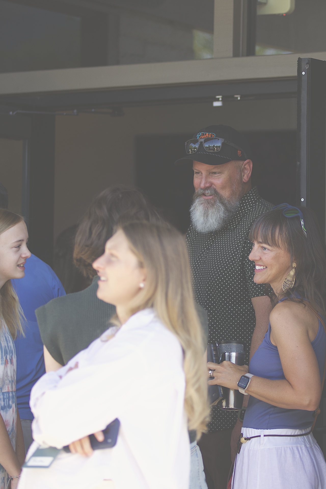 People socializing at an outdoor event, including a man with a gray beard and sunglasses on his cap, wearing a black and white polka dot shirt, and a woman with dark hair and a blue top, smiling while holding a drink.