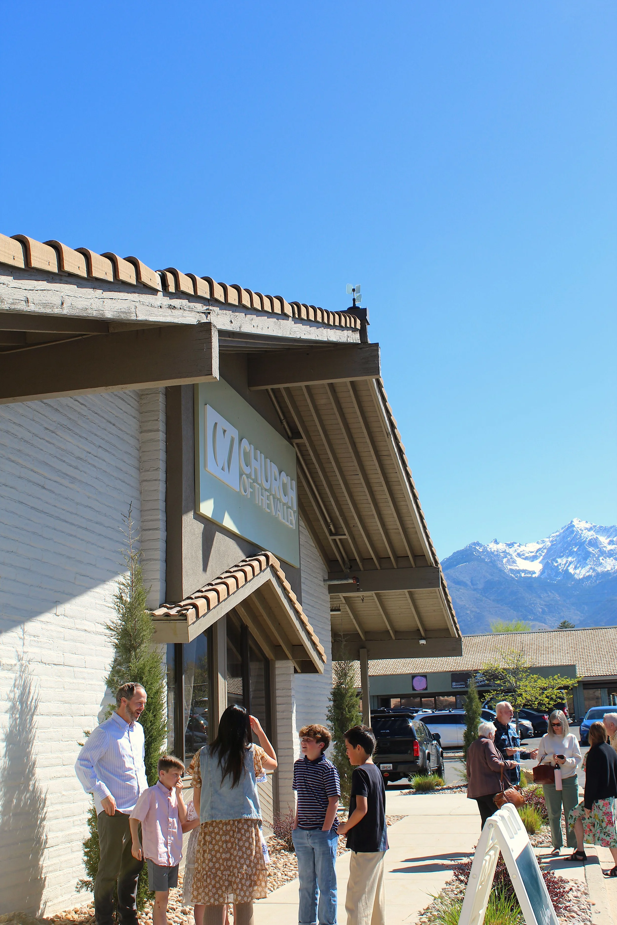 People standing outside a building with a sign that reads 'Church of the Valley', under a clear blue sky with snow-capped mountains in the background, during daytime.