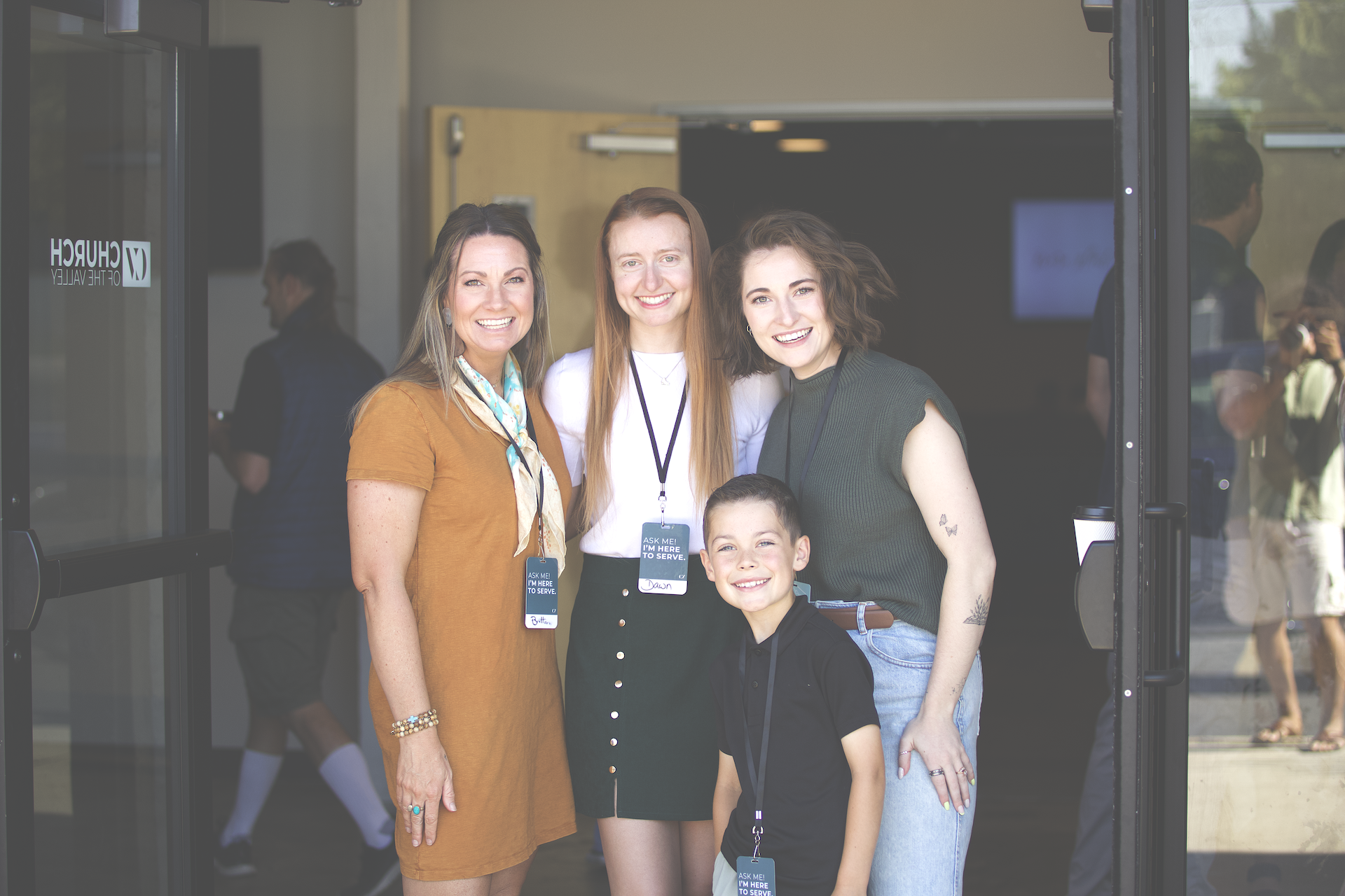 Four women and two boys smiling and standing in front of an open door at an event. The women are wearing badges that say they are there to serve, and the boys are smiling in front of them. The background shows other people at the event outside.