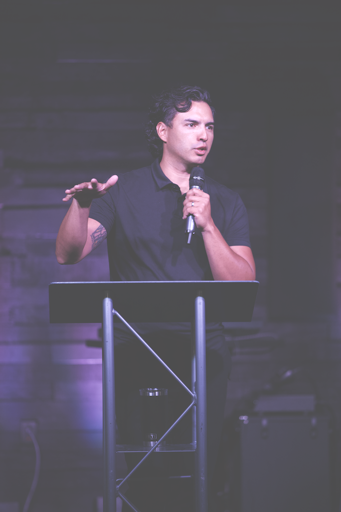 A man with dark, curly hair speaking into a microphone behind a black podium, gesturing with his left hand, in a dimly lit room with wood paneling.