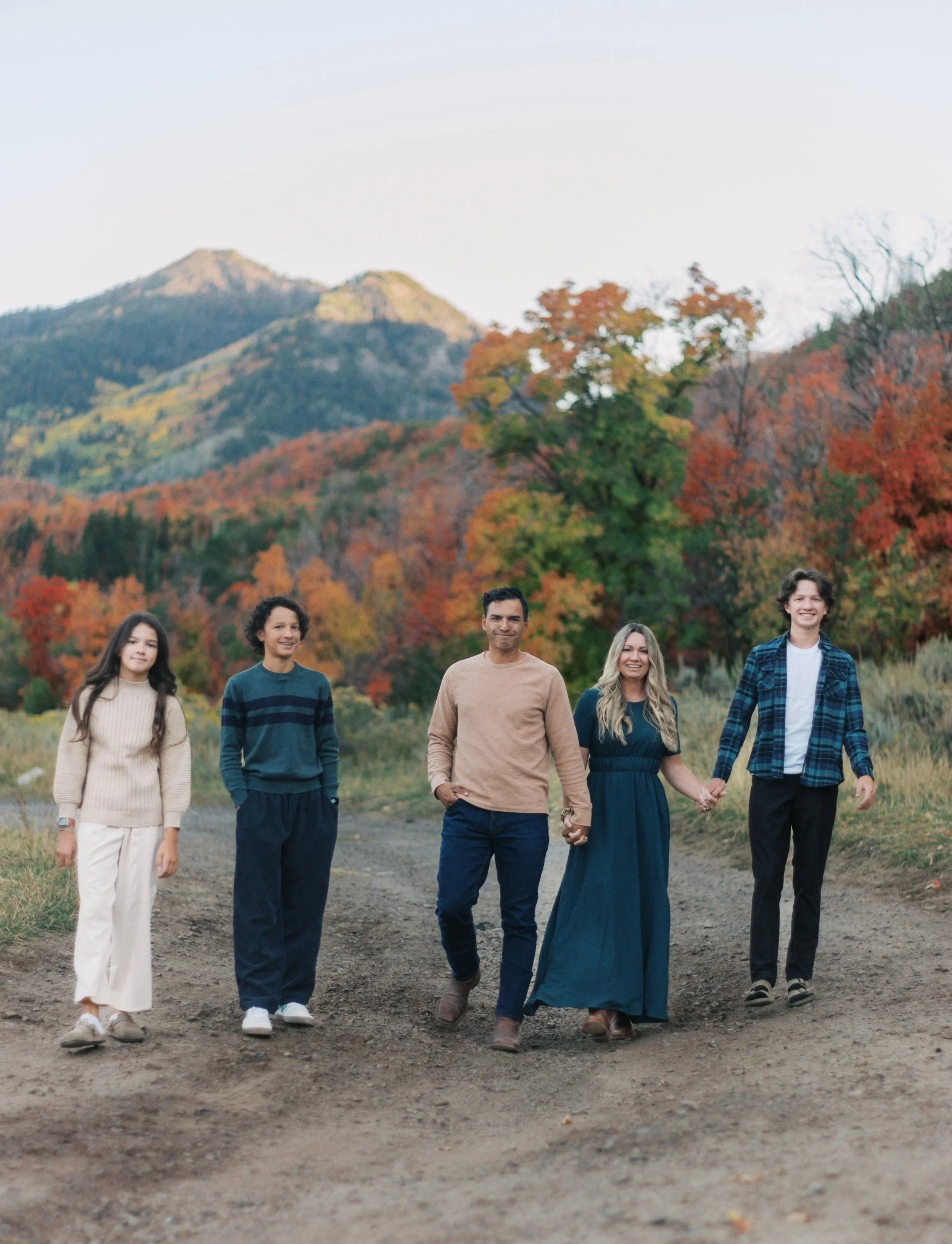 A group of six diverse people walking on a dirt path in a park with colorful autumn trees and mountains in the background, smiling and holding hands.