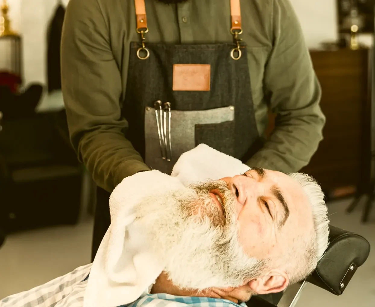 A barber giving a shave to an elderly man with a white beard at a barbershop.