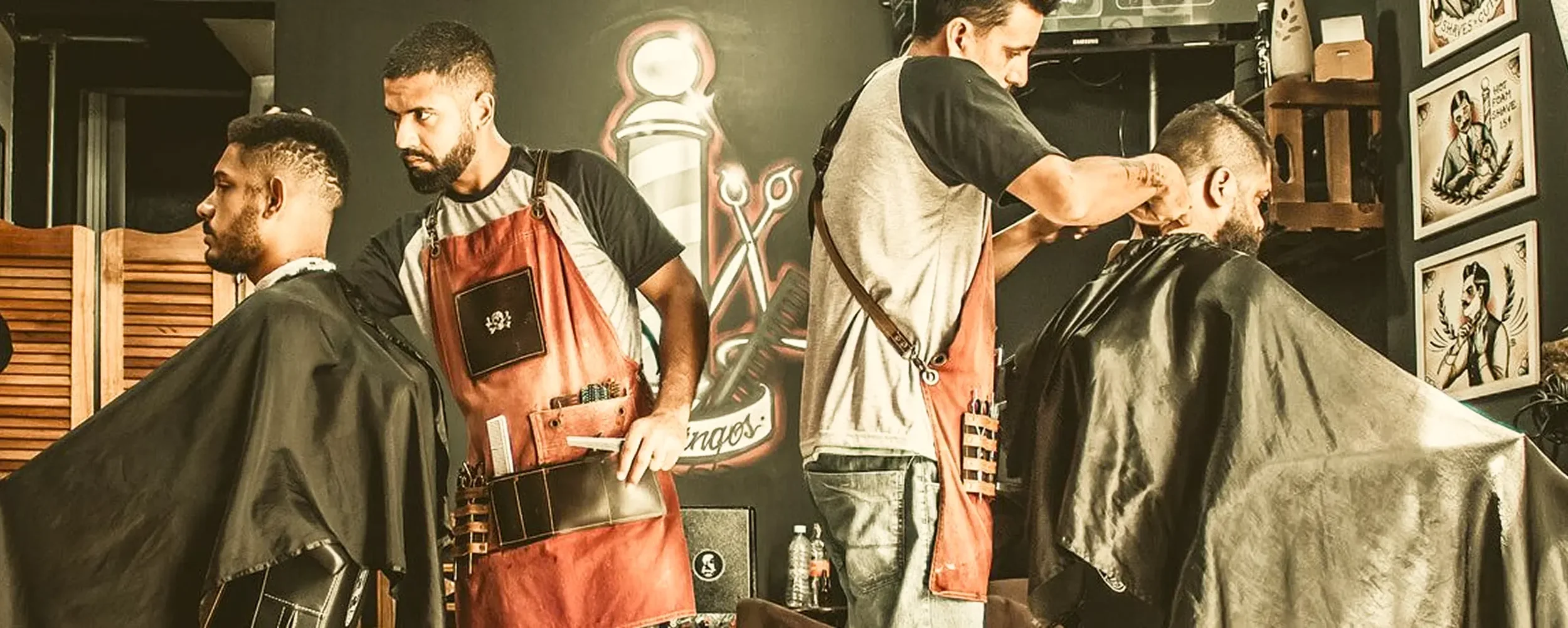 Barbershop scene with two barbers cutting men's hair, one with a beard in a gray shirt, the other with dark hair in a black shirt, and two clients seated in barber chairs with black capes. Barbers are focusing on their work, with framed artwork on th