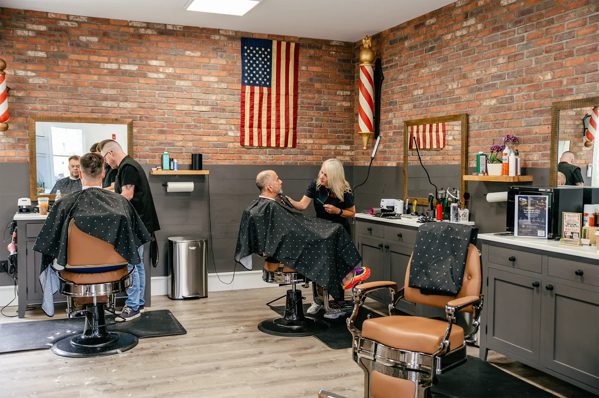 Two barbers cut hair for seated clients in a barbershop with brick walls, mirrors, and an American flag on display.