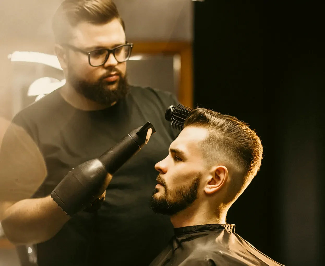Barber styling a man's hair in a salon with a blow dryer and round brush.
