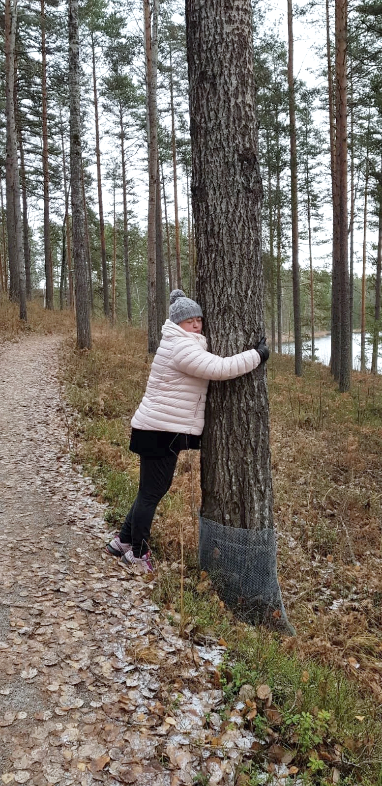 Nainen halaamassa puuta metsätiellä, taustalla puita ja järvi, syksyinen ilma, päällä pipo ja lämmin takki.