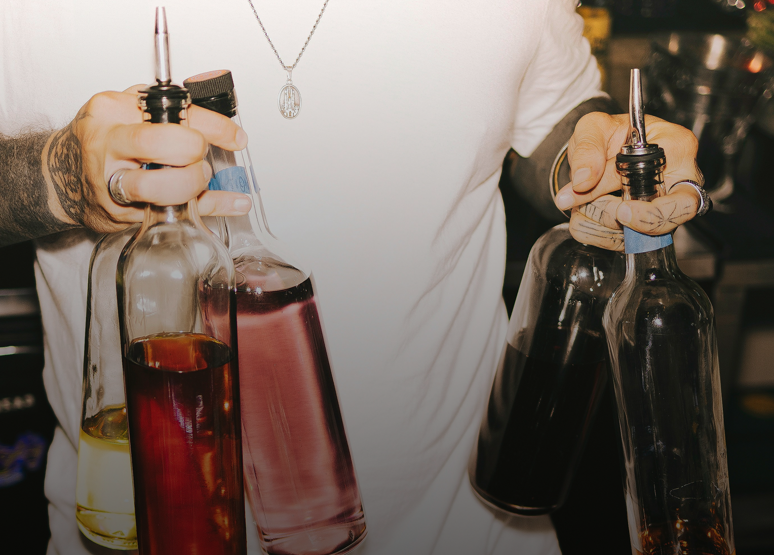 Person holding bottles of different colored liquids, using a pour spout, in a kitchen or bar setting.