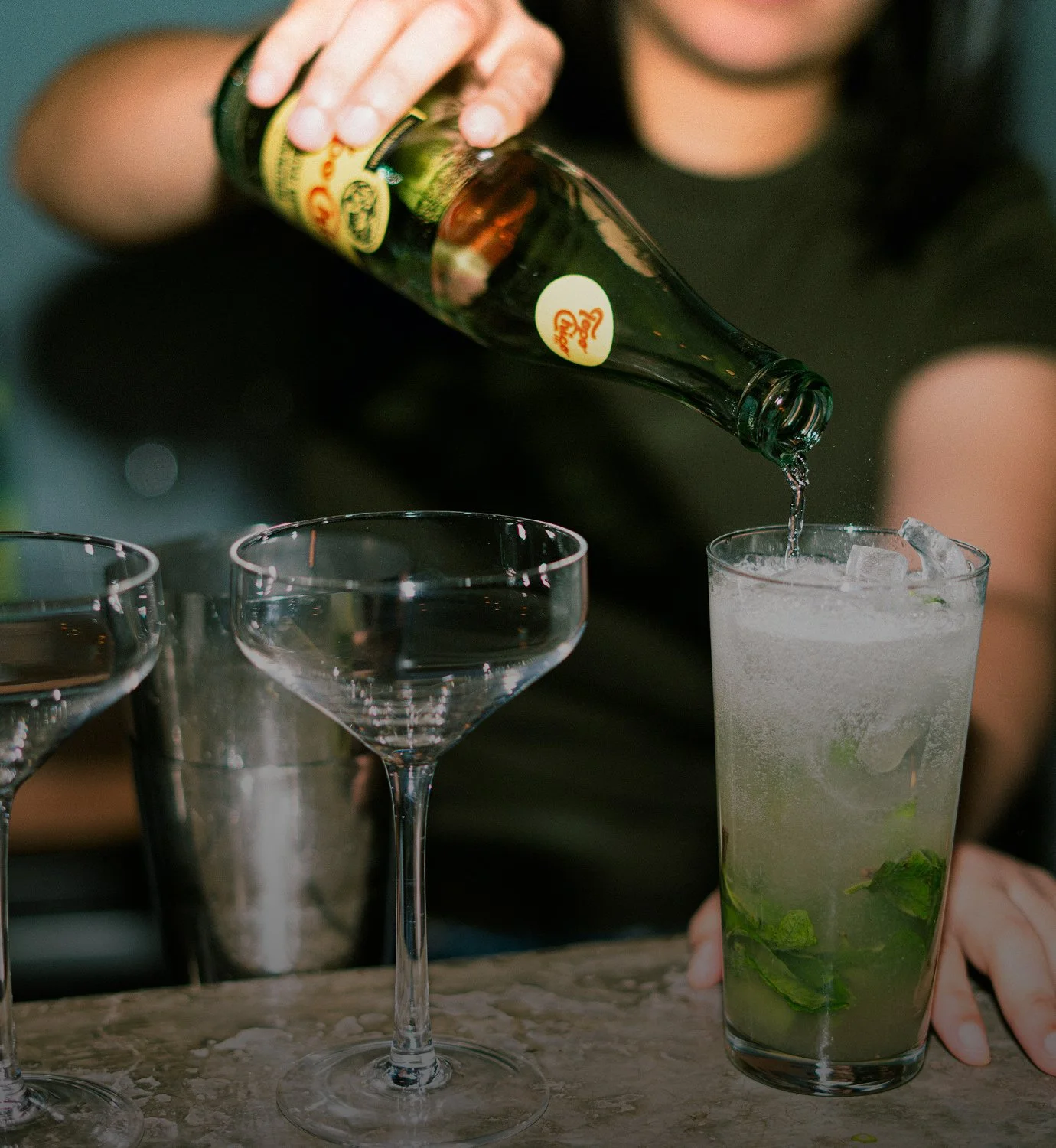 A person pouring a drink from a whiskey bottle into a tall glass with ice and mint leaves, with other cocktail glasses on the table.