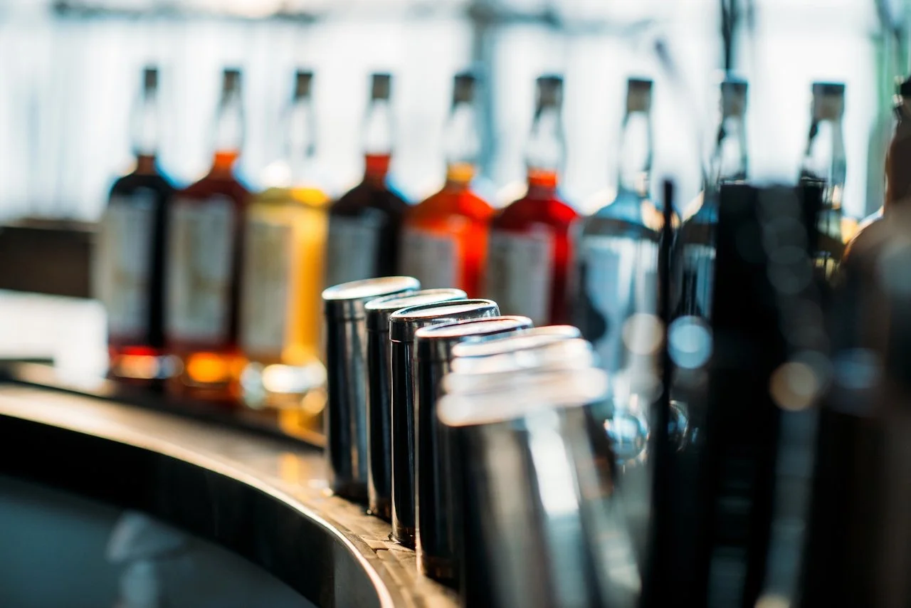 A row of bottle dispensers with metal shot glasses in front, lined up on a counter, with blurred colorful bottles in the background.
