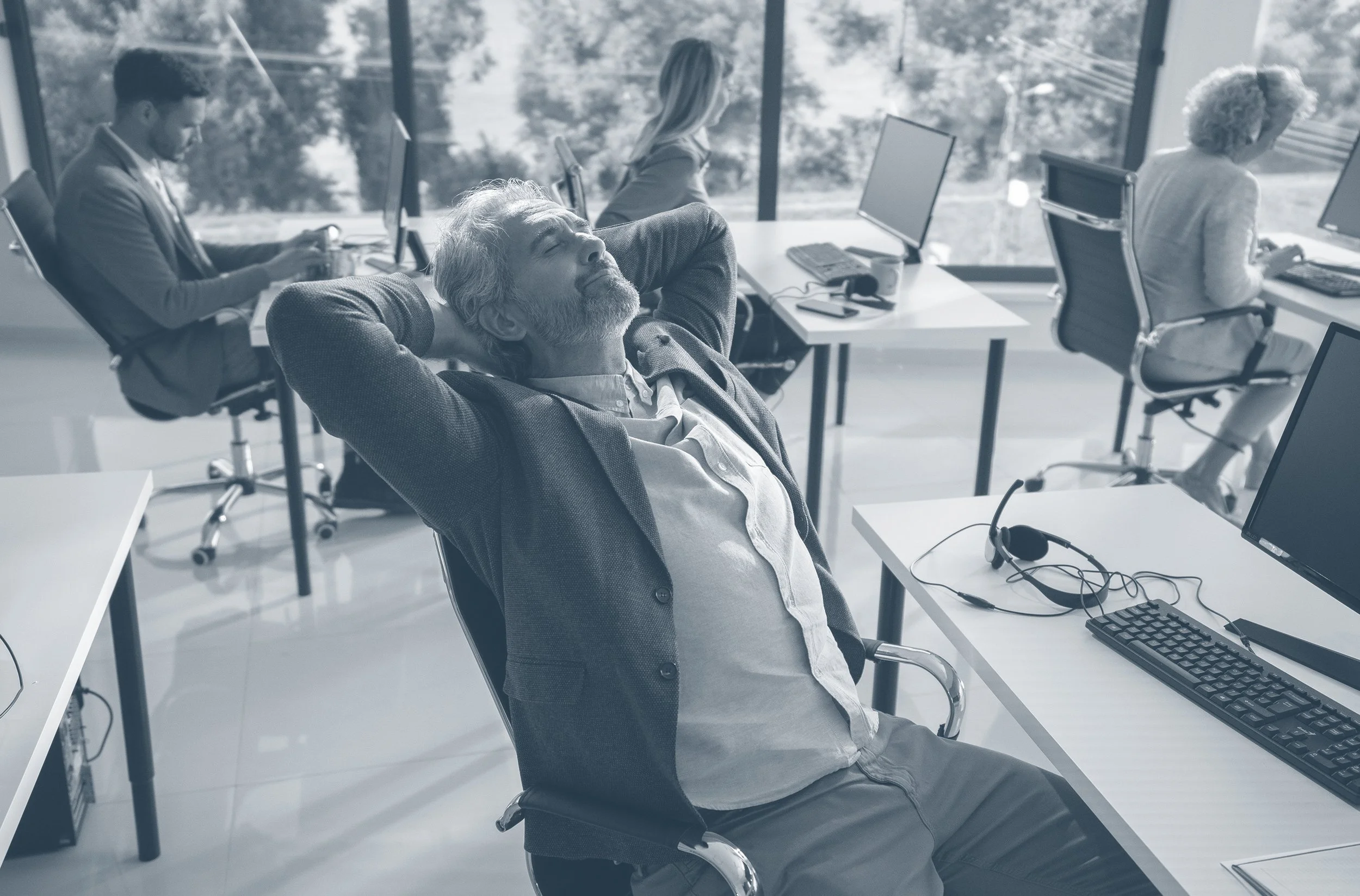 Man relaxing with his hands behind his head at a modern office, with other employees working at their desks in the background, near large windows showing trees outside.