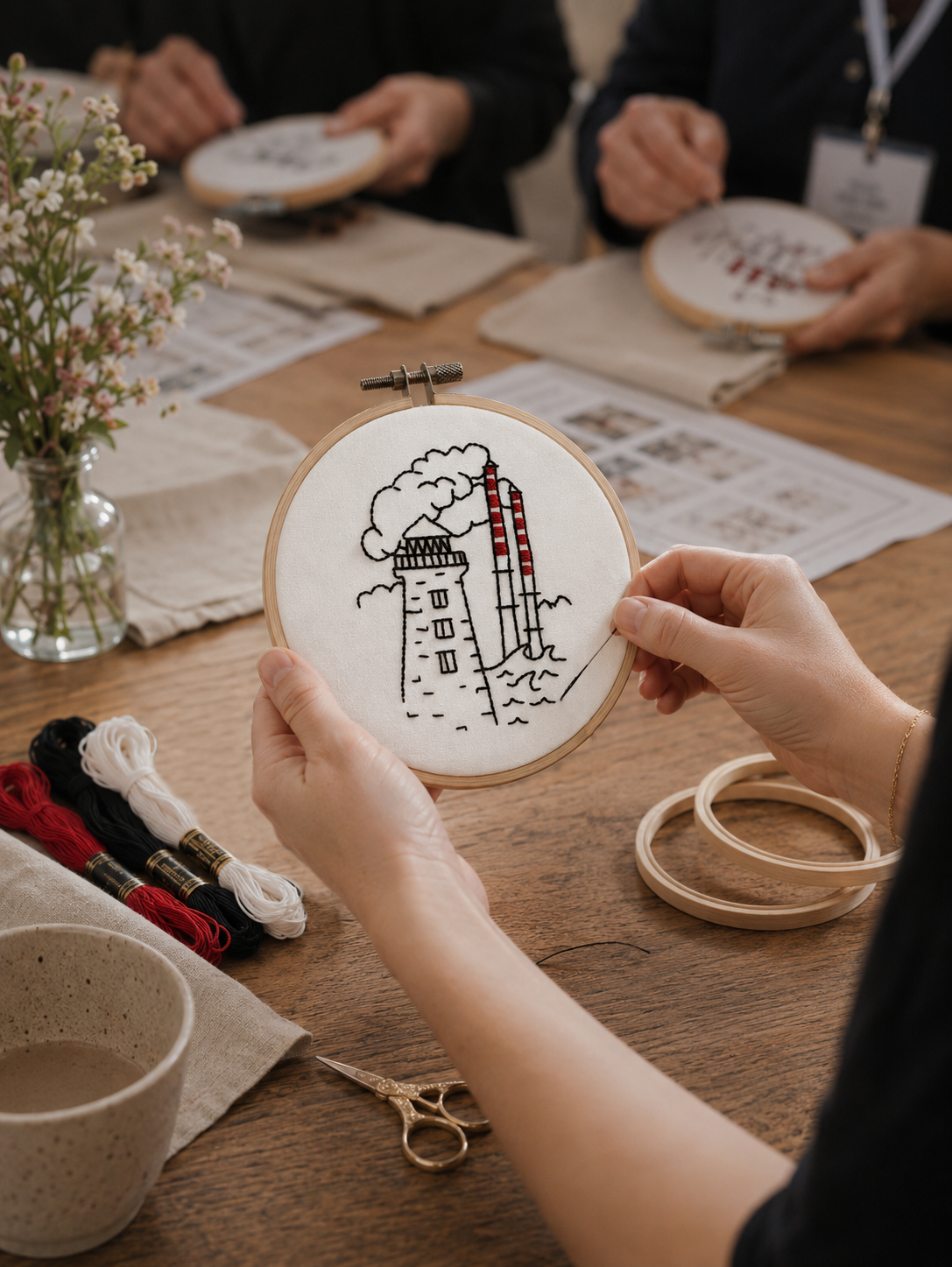 Person holding an embroidery hoop with a factory smoke stack and chimneys embroidered on fabric, with crafting supplies on a wooden table.
