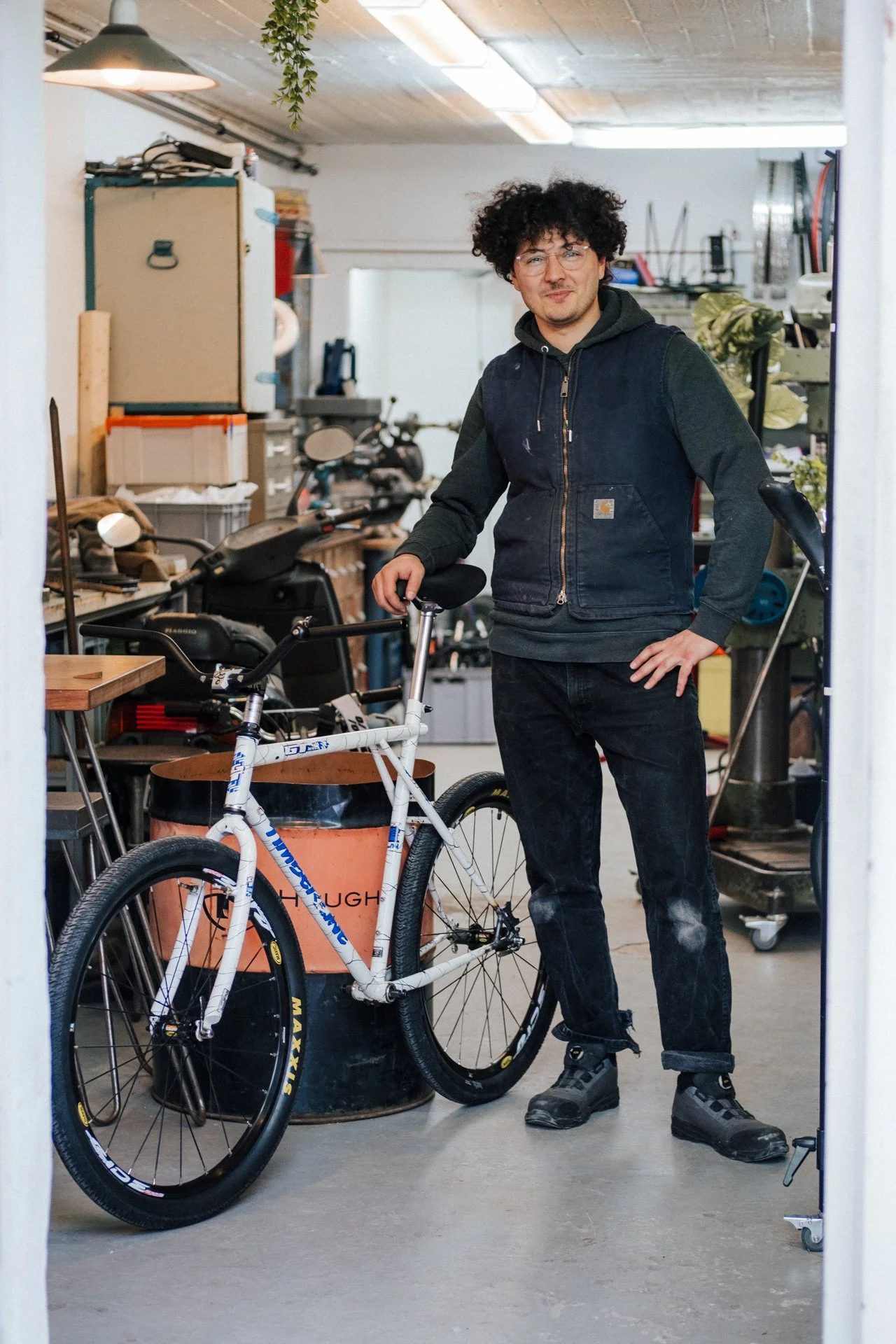 Ein junger Mann mit lockigem Haar, Brille und dunkler Kleidung steht in einer Werkstatt neben einem weißen Fahrrad mit schwarzen Reifen.