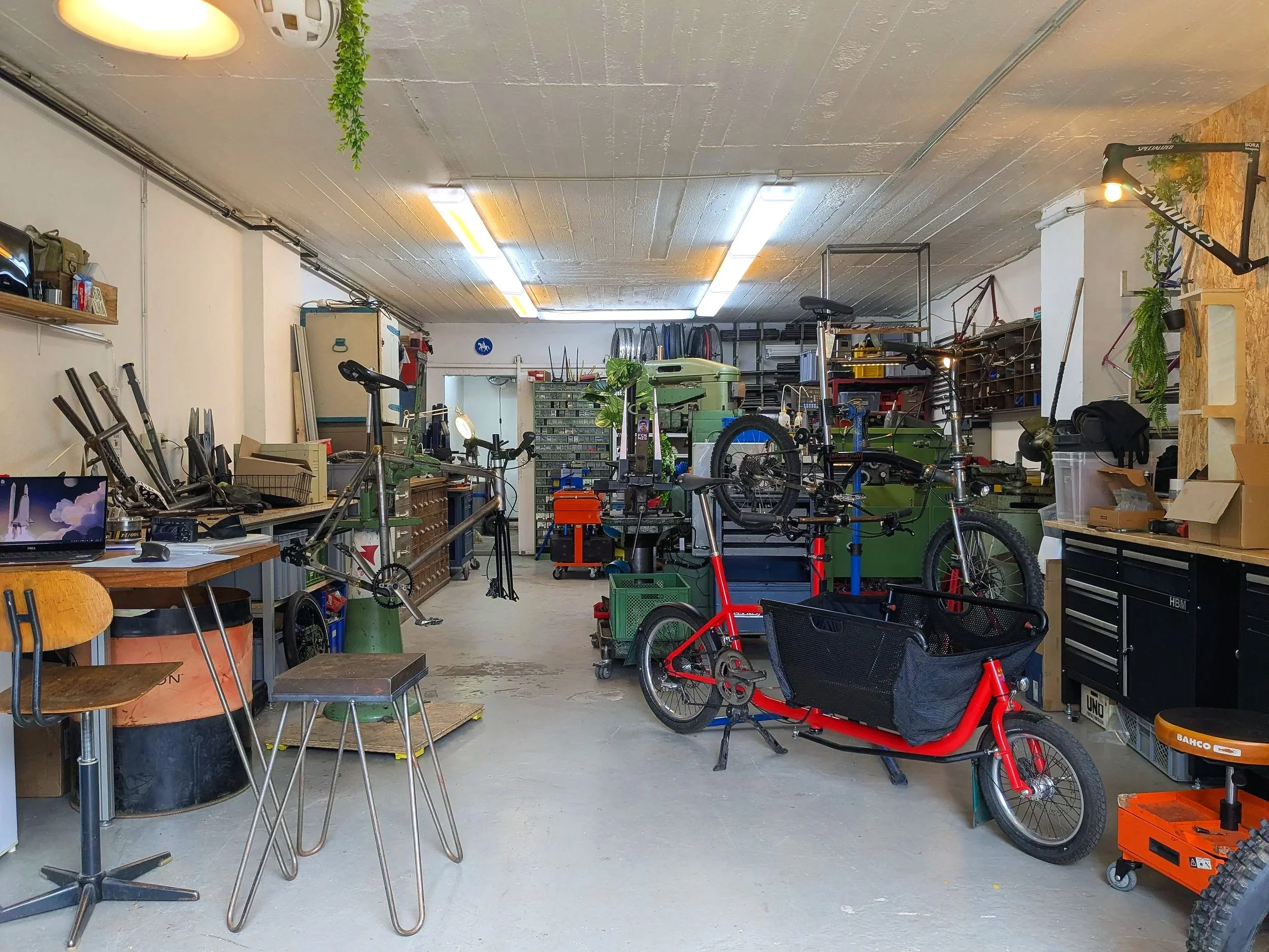 Ein Fahrradwerkstatt mit mehreren Fahrrädern, Werkzeugen und Werkbänken, einer Beleuchtung an der Decke und einigen Pflanzen an der Wand.