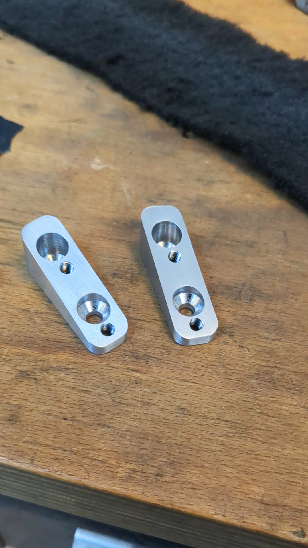 Zwei silberne Metallteile mit gebohrten Löchern auf einem Holzarbeitsbrett.