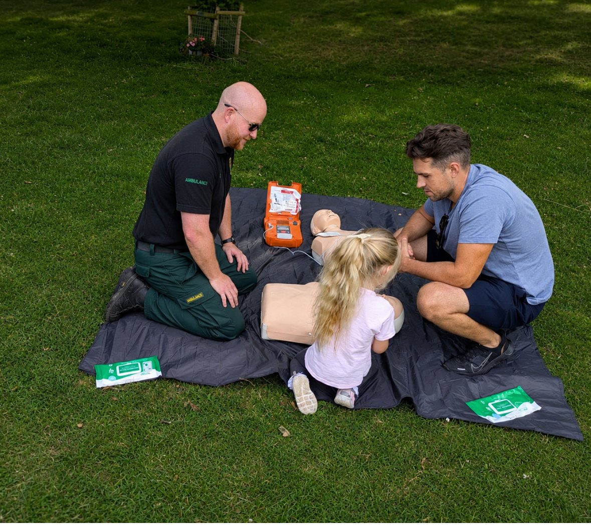 Children and adults practicing CPR on mannequins during an outdoor training session on a grassy field.