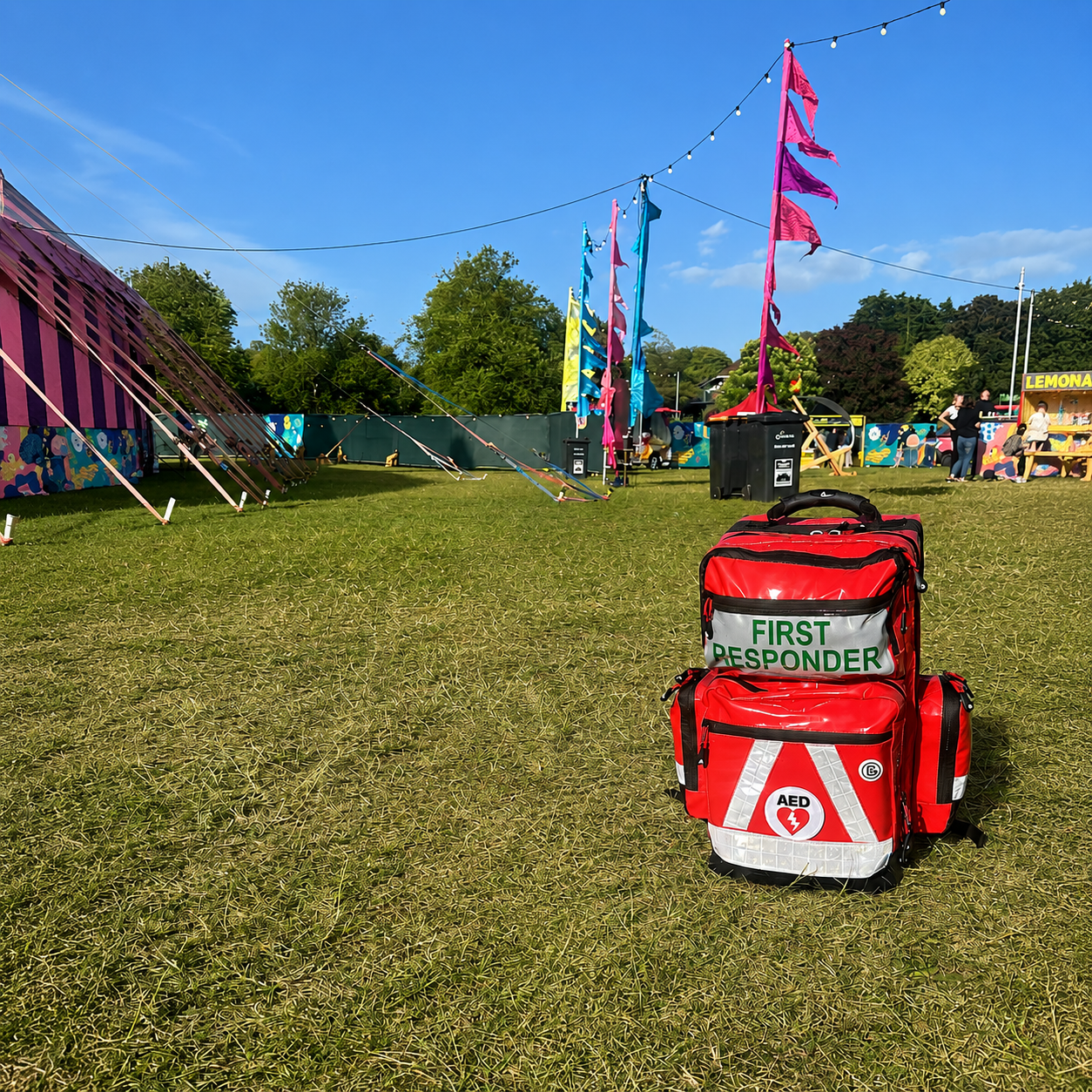 A first responder medical bag with AED symbol on grass at outdoor festival with colorful flags and tents in the background.