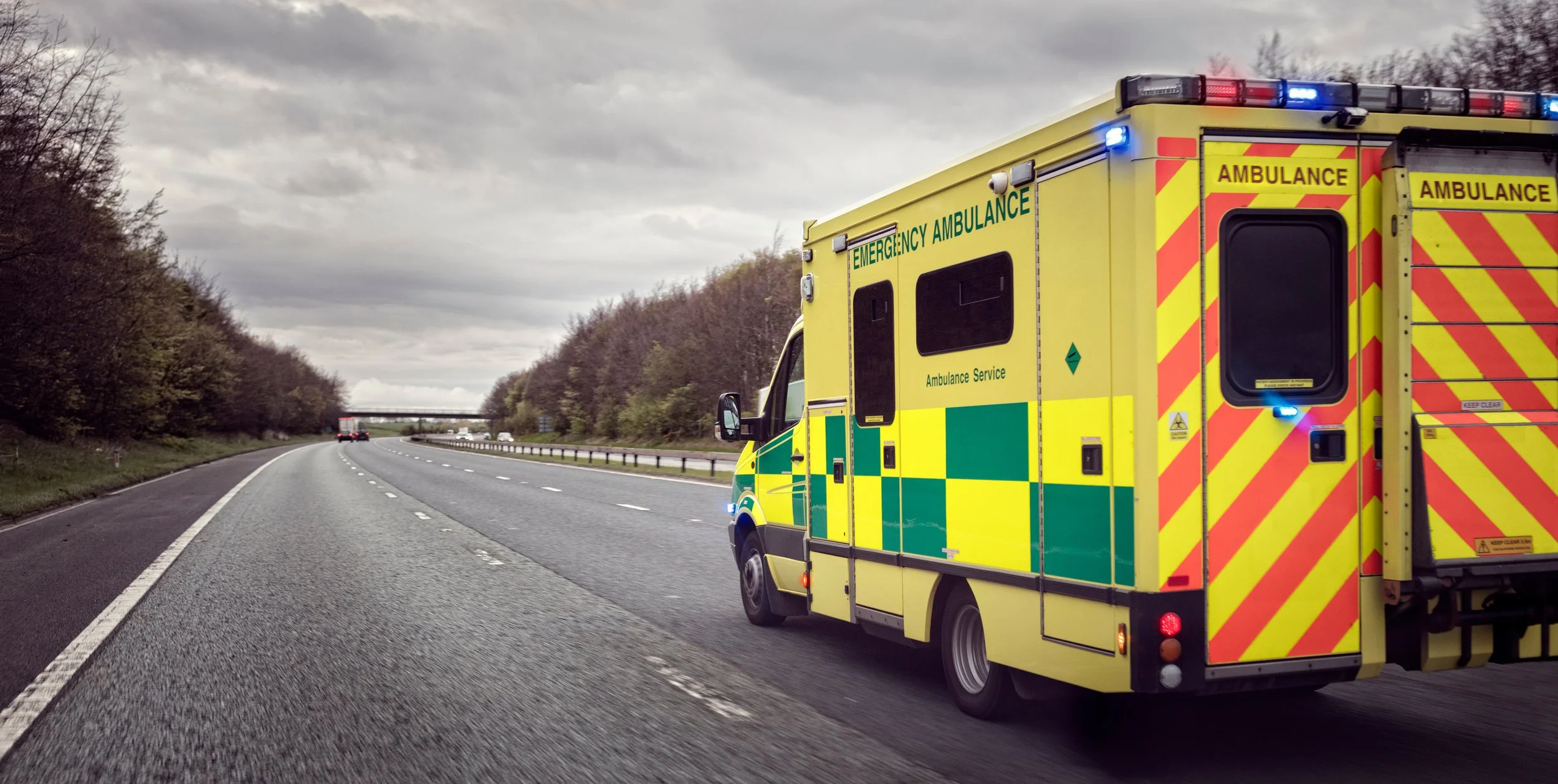 Yellow emergency ambulance on a highway, moving away, with trees on both sides and a cloudy sky.