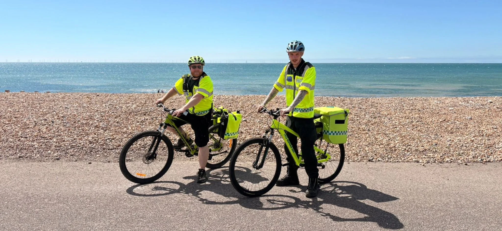 Two ambulance staff in high-visibility yellow jackets riding bicycles along a coastal road, with the beach and ocean in the background.