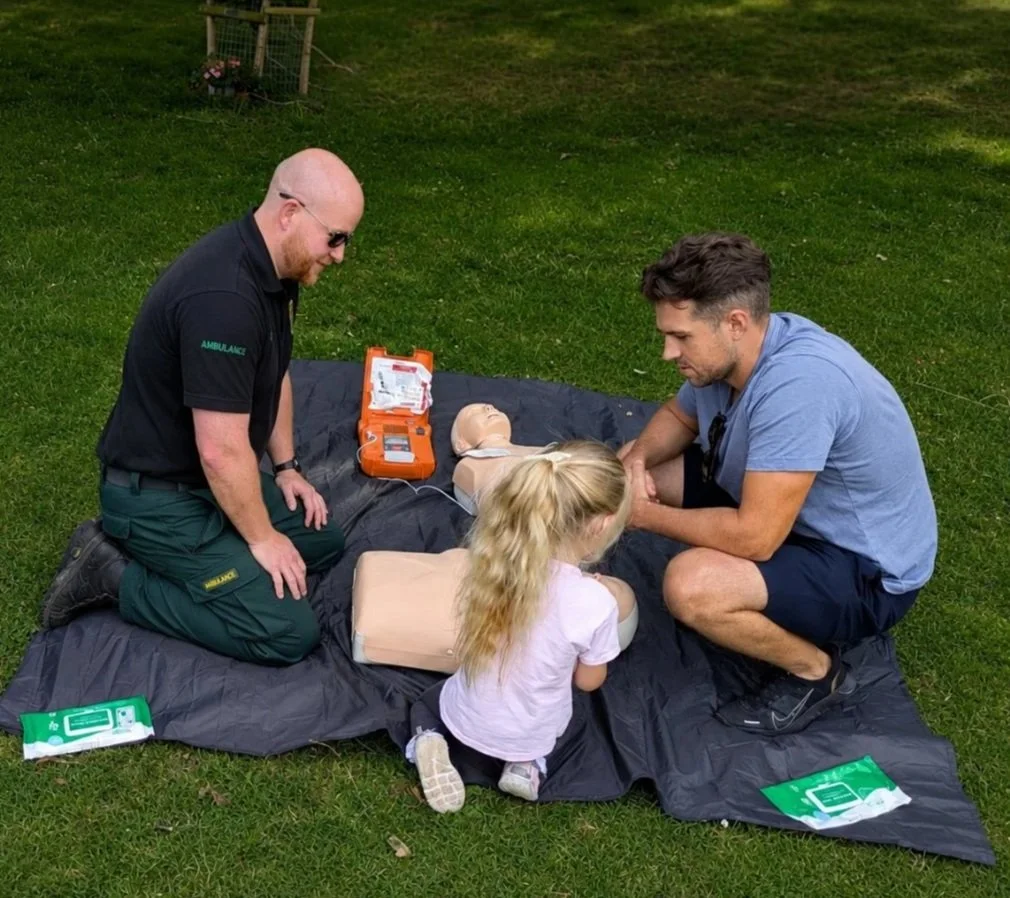 Two adults and a young girl practicing CPR on a training dummy outdoors on a black mat, with CPR training supplies nearby.