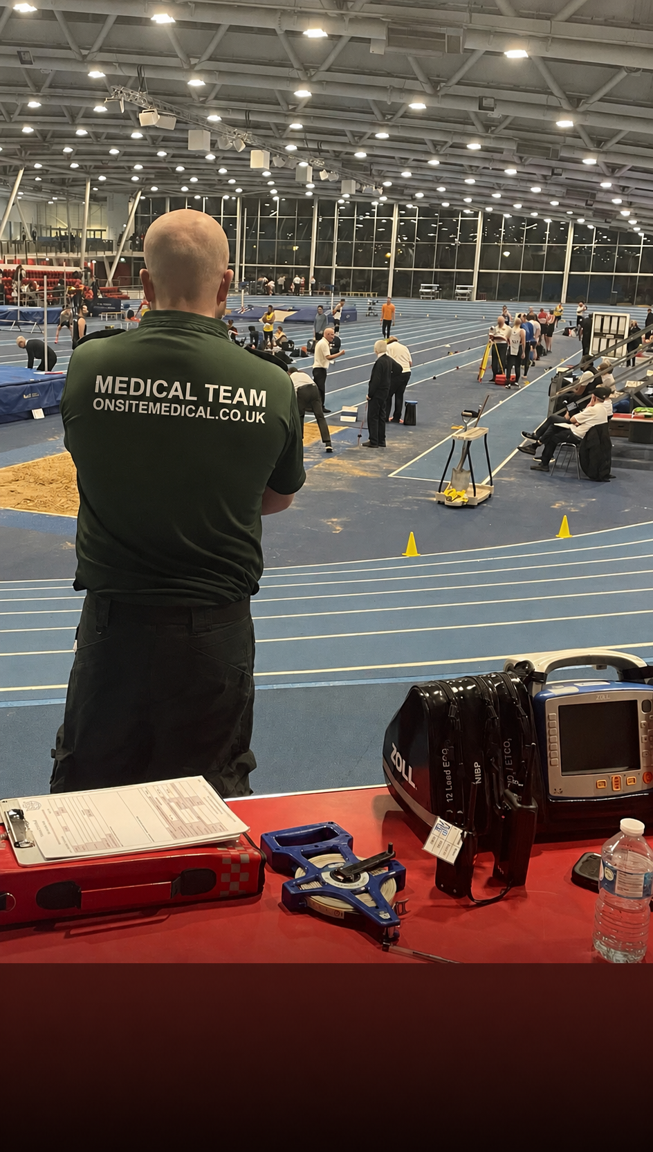 An indoor athletics arena with athletes competing in a track event, medical staff on scene, and a medical team member observing from behind in a green shirt labeled 'Medical Team'.