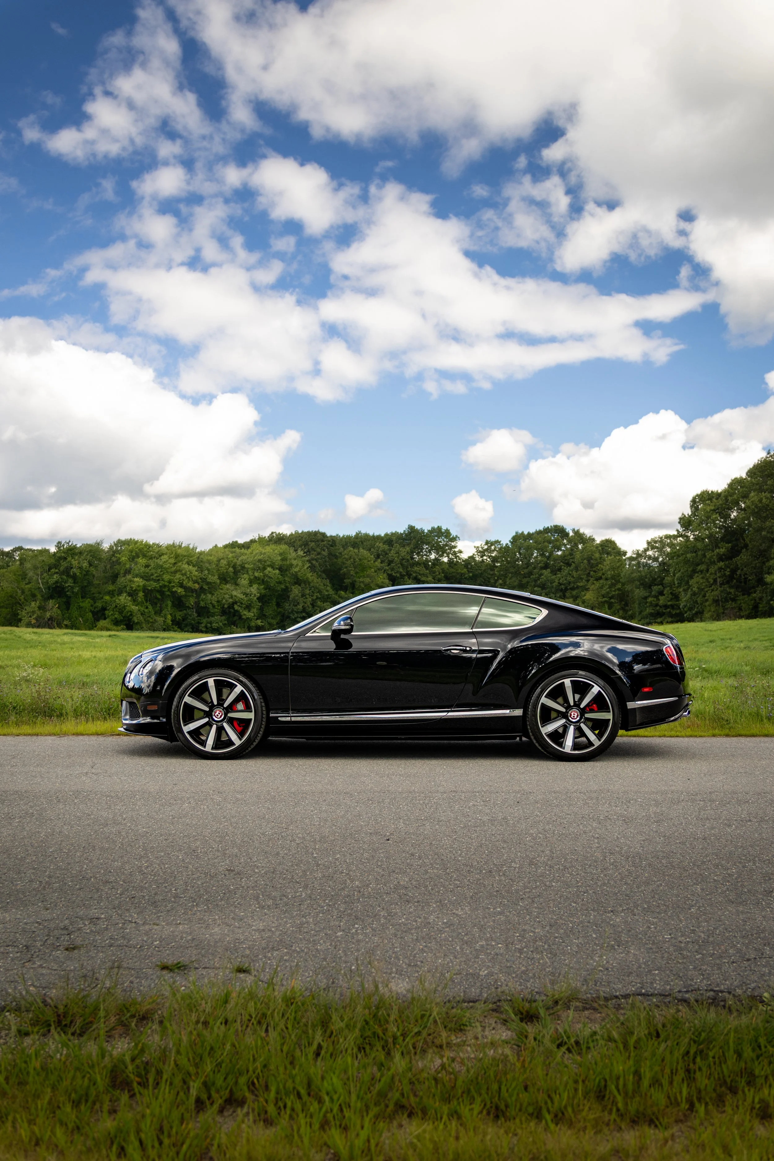 Bentley Continental GT V8 S parked on a road with a grassy field in the background and cloudy blue sky.