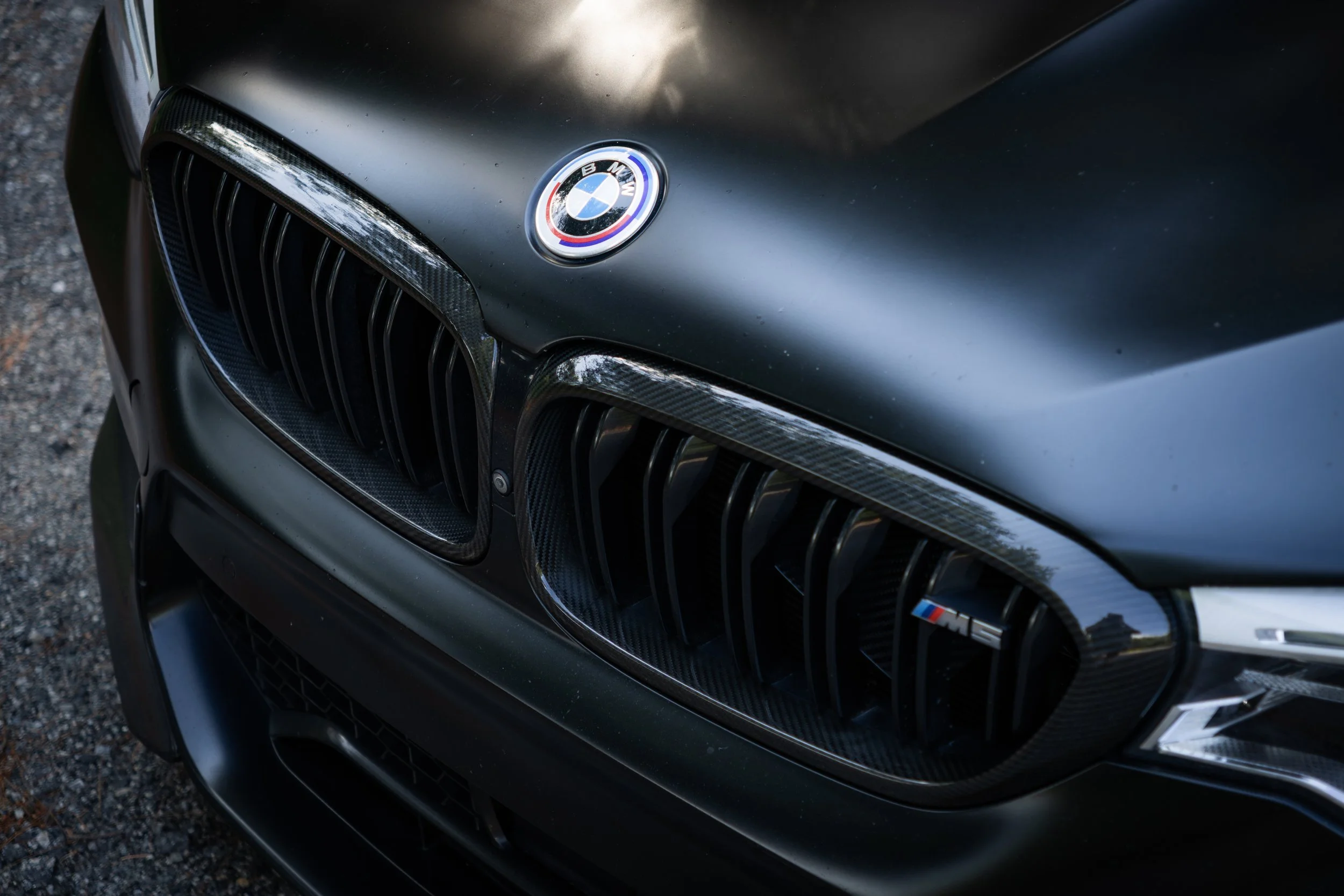 Close-up of the front of a flat black BMW M5 Competition, showing the BMW emblem on the hood and part of the carbon fiber front grille.