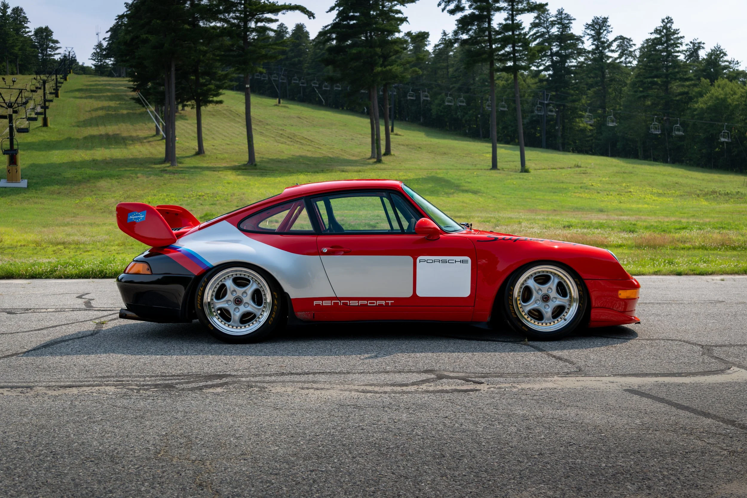 A red and white Porsche GT2 Supercup race car with black, gray, and blue accents parked on a paved surface, with a grassy hillside and ski lift chairs in the background.
