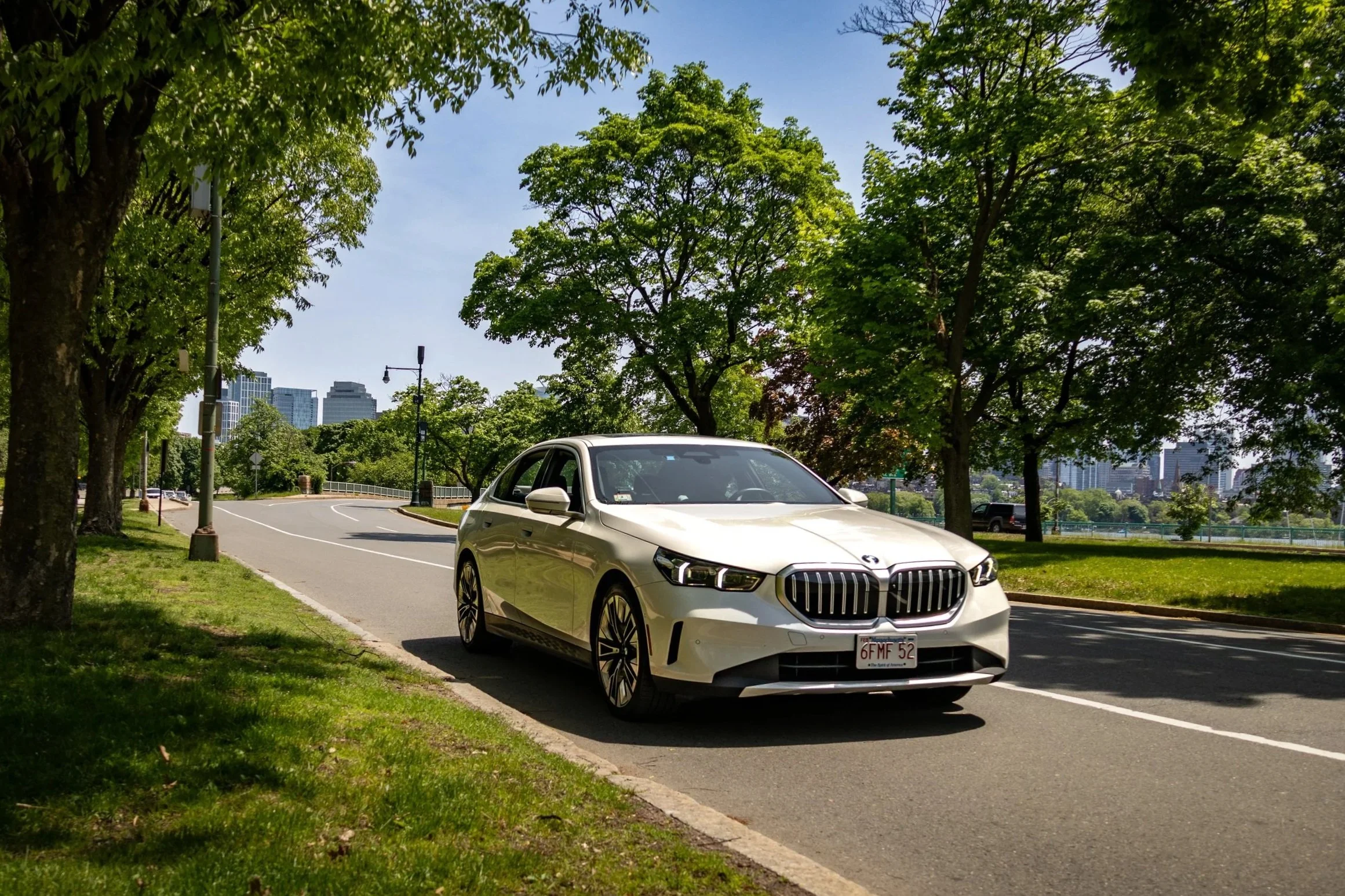 A white BMW 5-series test vehicle, owned by the MIT Age Lab parked on the side of a street under green leafy trees with a city skyline in the background.