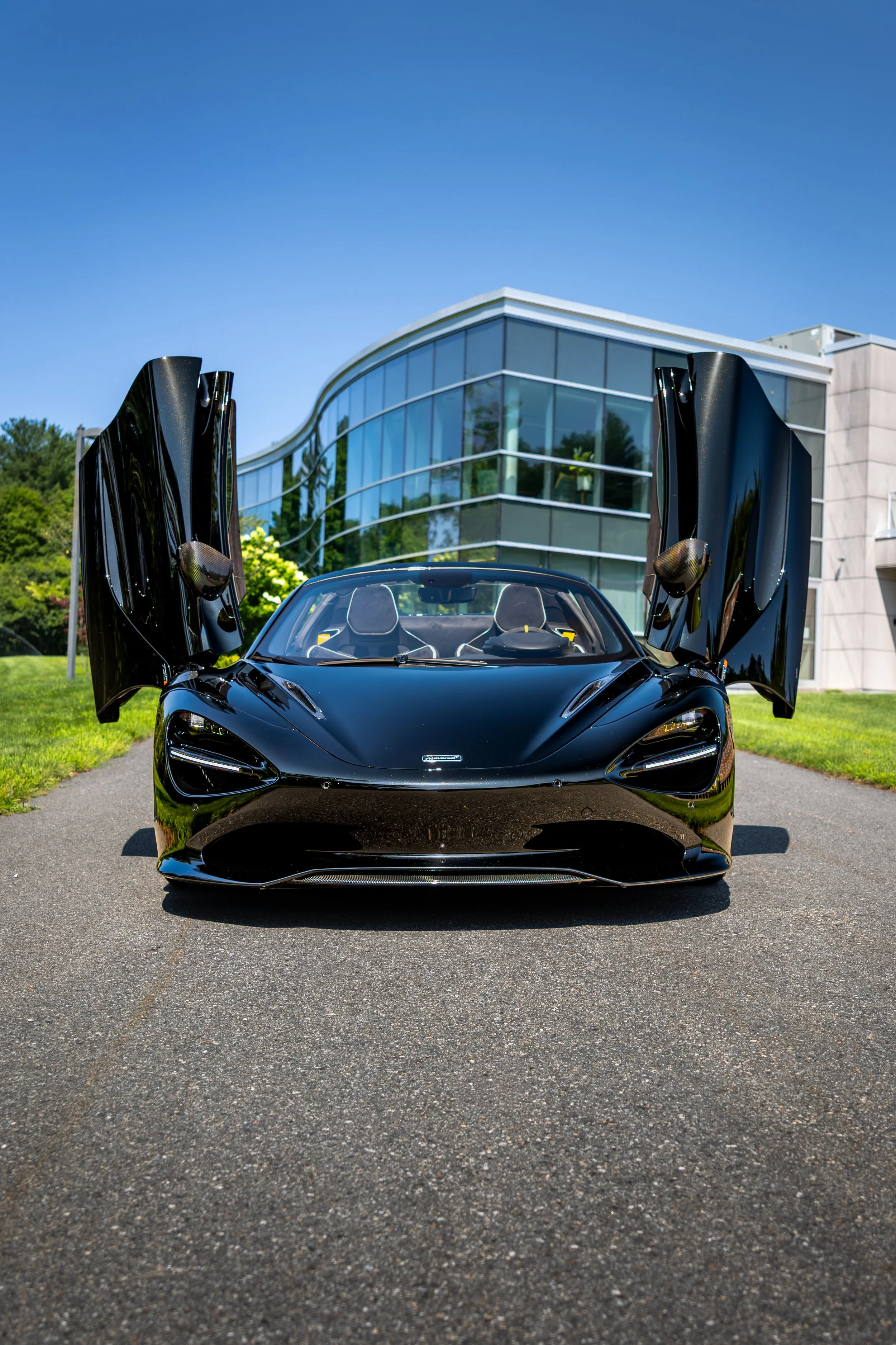 A black McLaren 750s with its butterfly doors open, parked in front of a modern glass building on a sunny day.