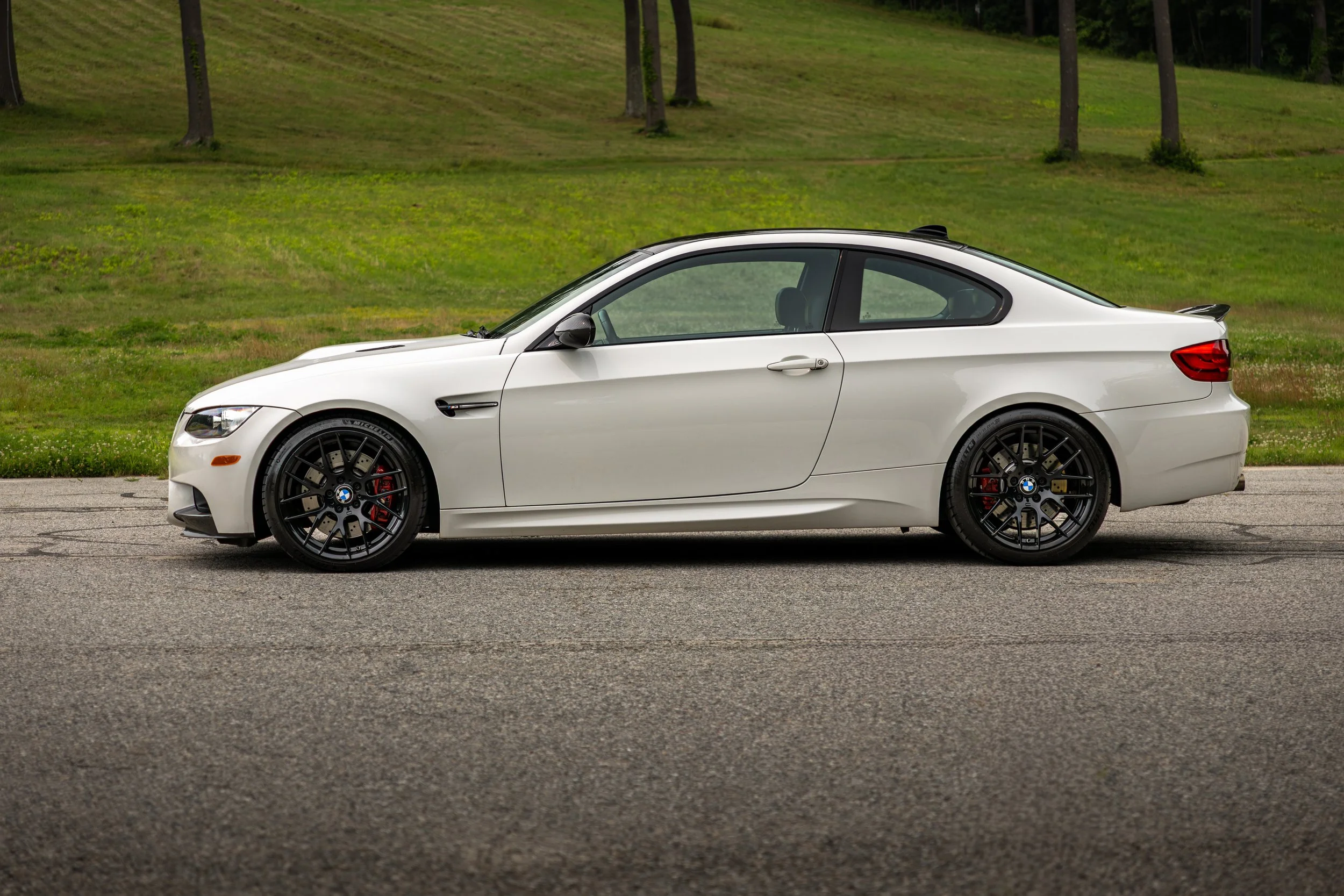White BMW M3 coupe parked on a paved surface with grassy hills and trees in the background.