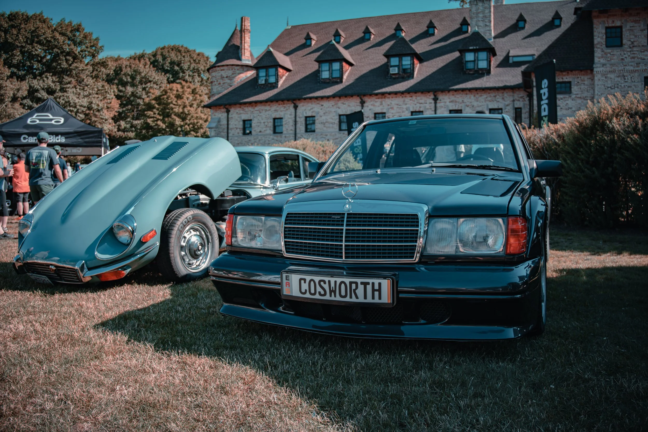 A vintage Jaguar with its hood open and a Black Mercedes 190 Cosworth with a European license plate that reads 'COSWORTH' are on display outdoors at the Larz Anderson Museum in Brookline, MA.