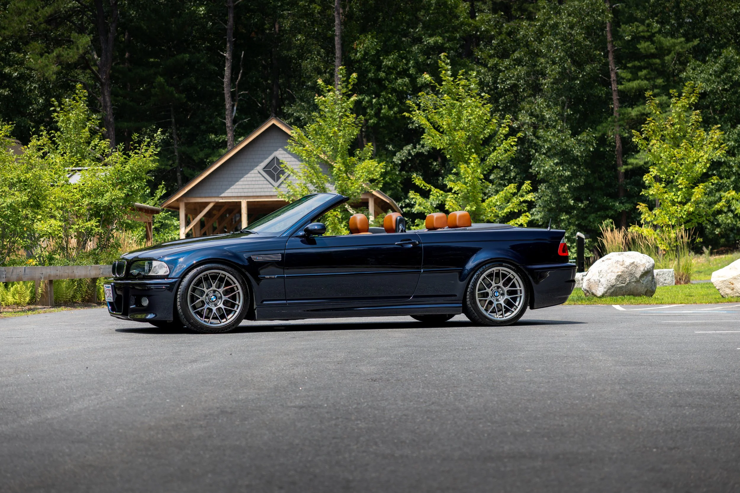 A dark blue E46 BMW M3 Convertible parked on a paved surface with the top down, surrounded by greenery and trees in the background.