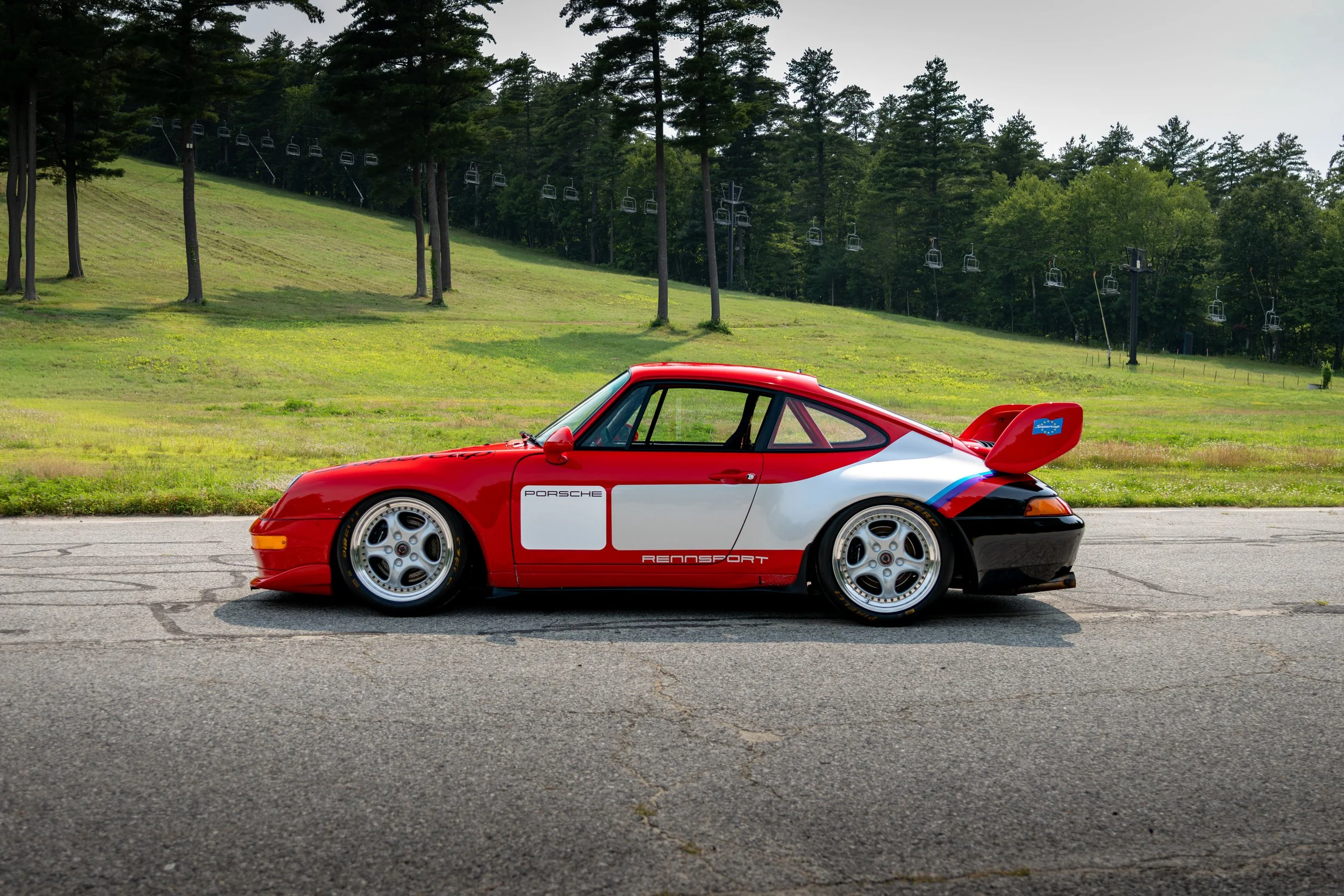 A red and white Porsche GT2 Supercup race car with racing decals parked on a paved road, with a green hillside and ski lift chairs in the background.
