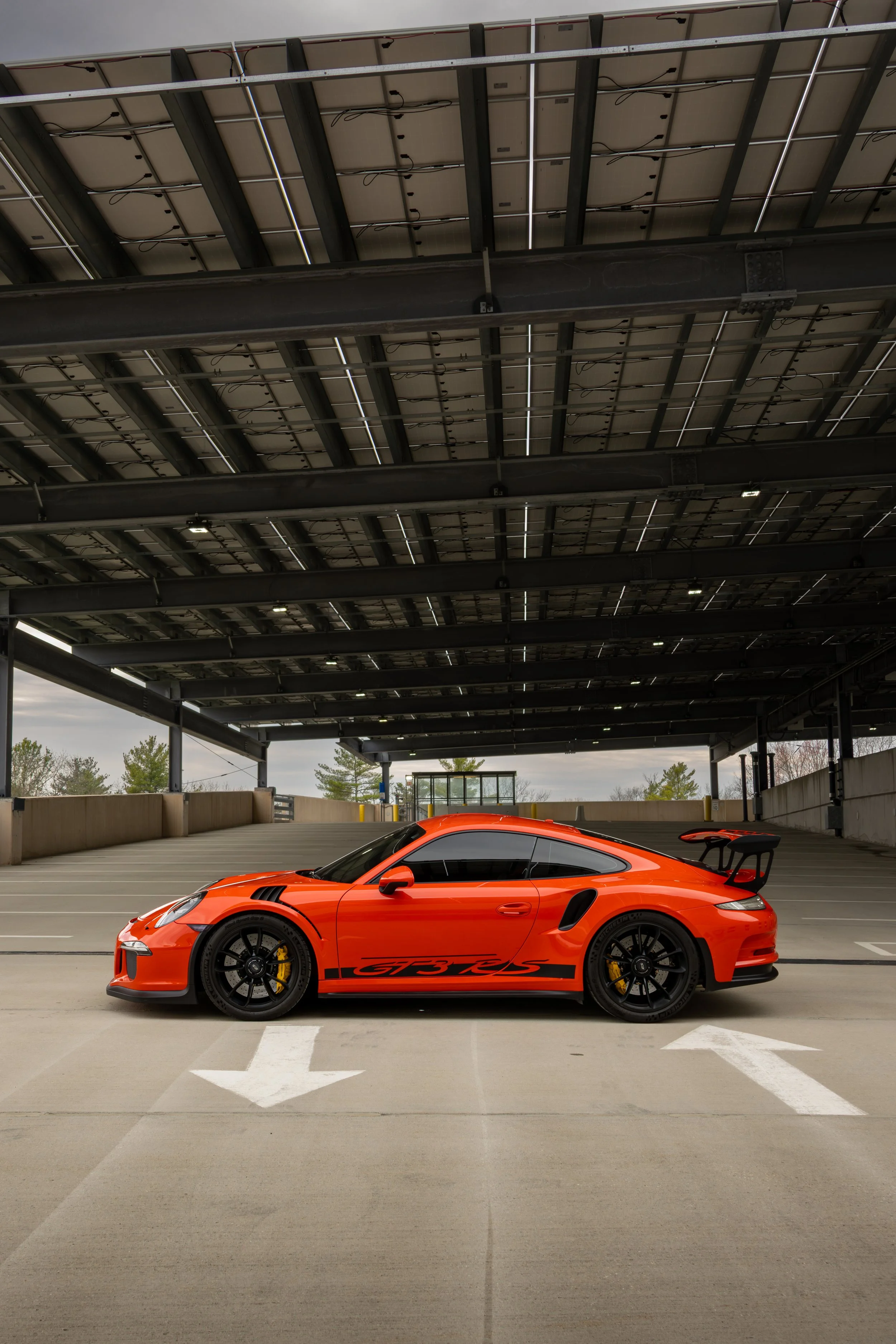 Lava orange Porsche 911 GT3 RS parked in an outdoor parking structure, facing left, with clouds overhead and some trees in the background.