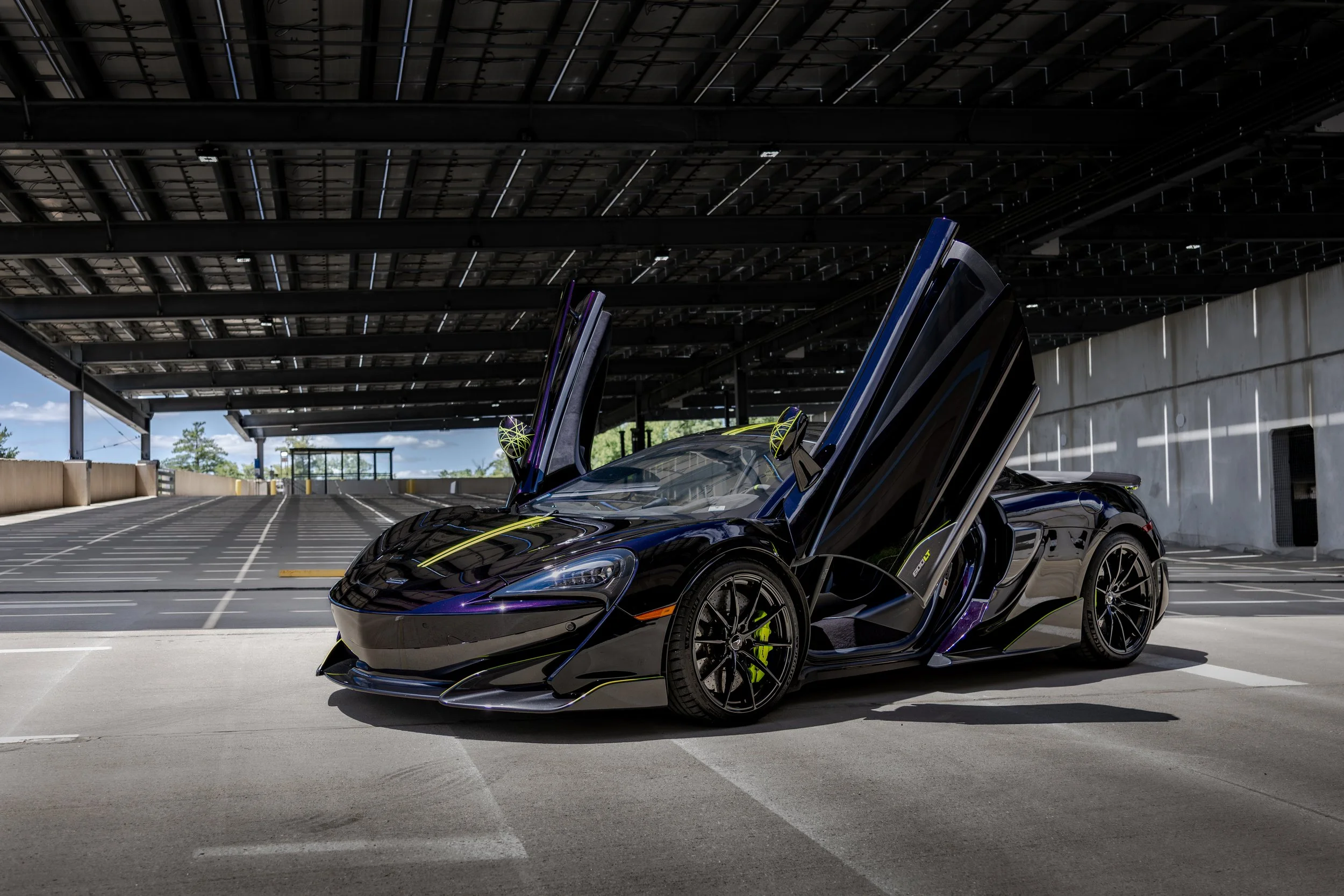 A McLaren 600LT special edition, 1 of 12, featuring lime green accents and gull-wing doors open, parked under a empty covered parking lot on a sunny day.