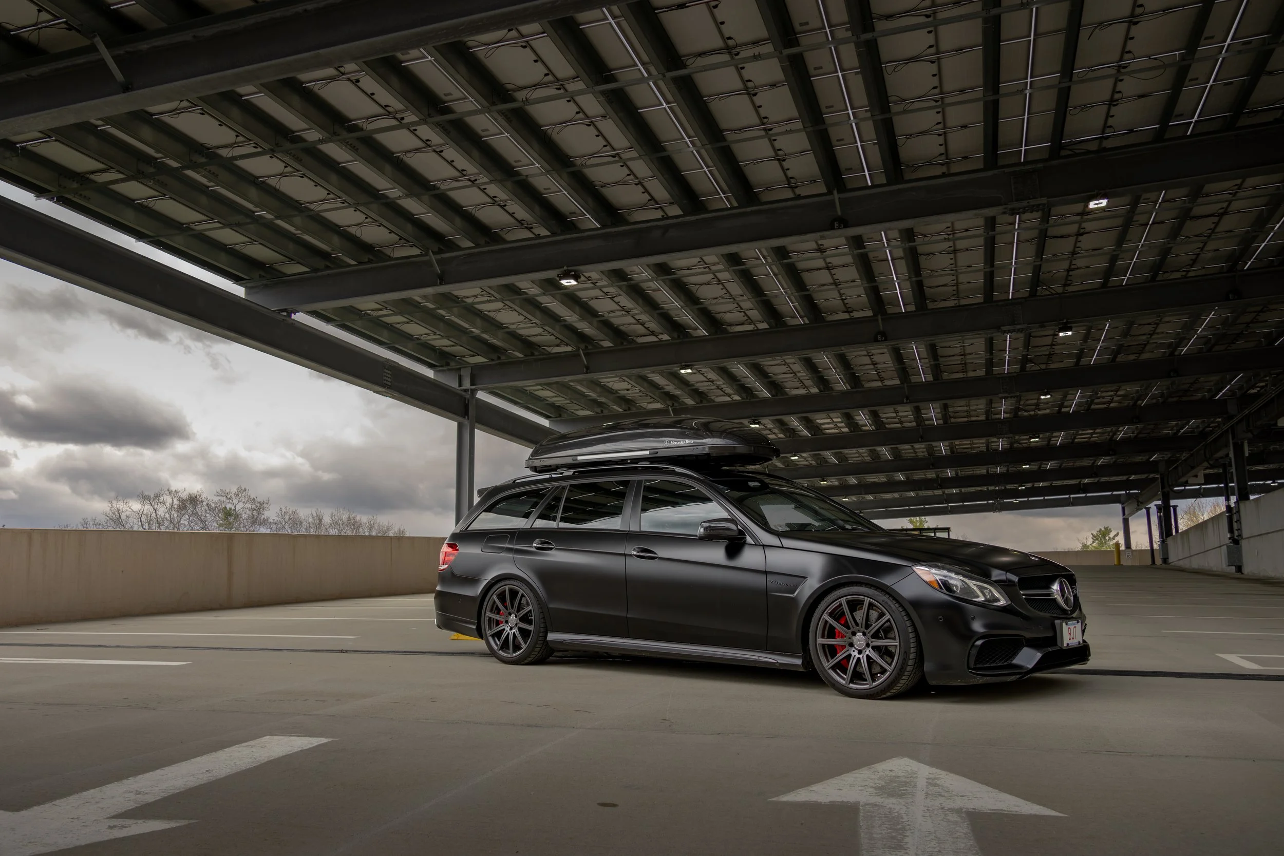 A lowered flat black 2015 Mercedes-Benz E63s AMG Wagon (S212) with a black roof box parked under a covered parking area, with cloudy sky in the background.