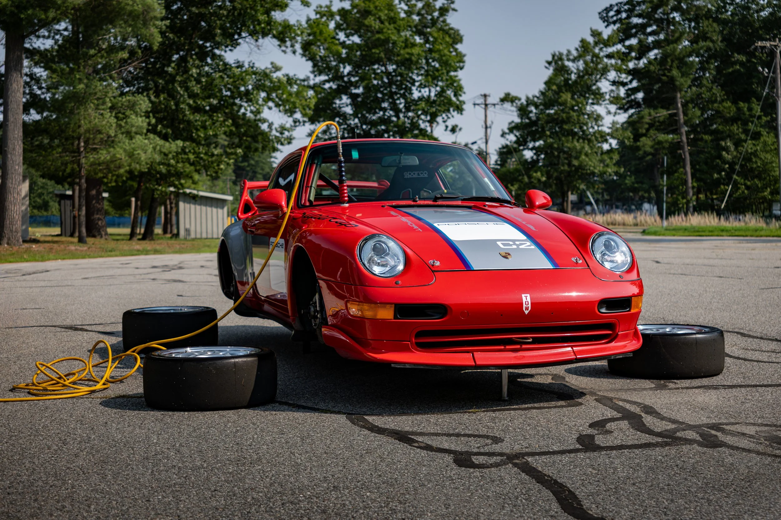 Red Porsche GT2 Supercup race car with racing stripes parked on pavement, raised on air jacks, surrounded by tires and an air hose with trees in the background.