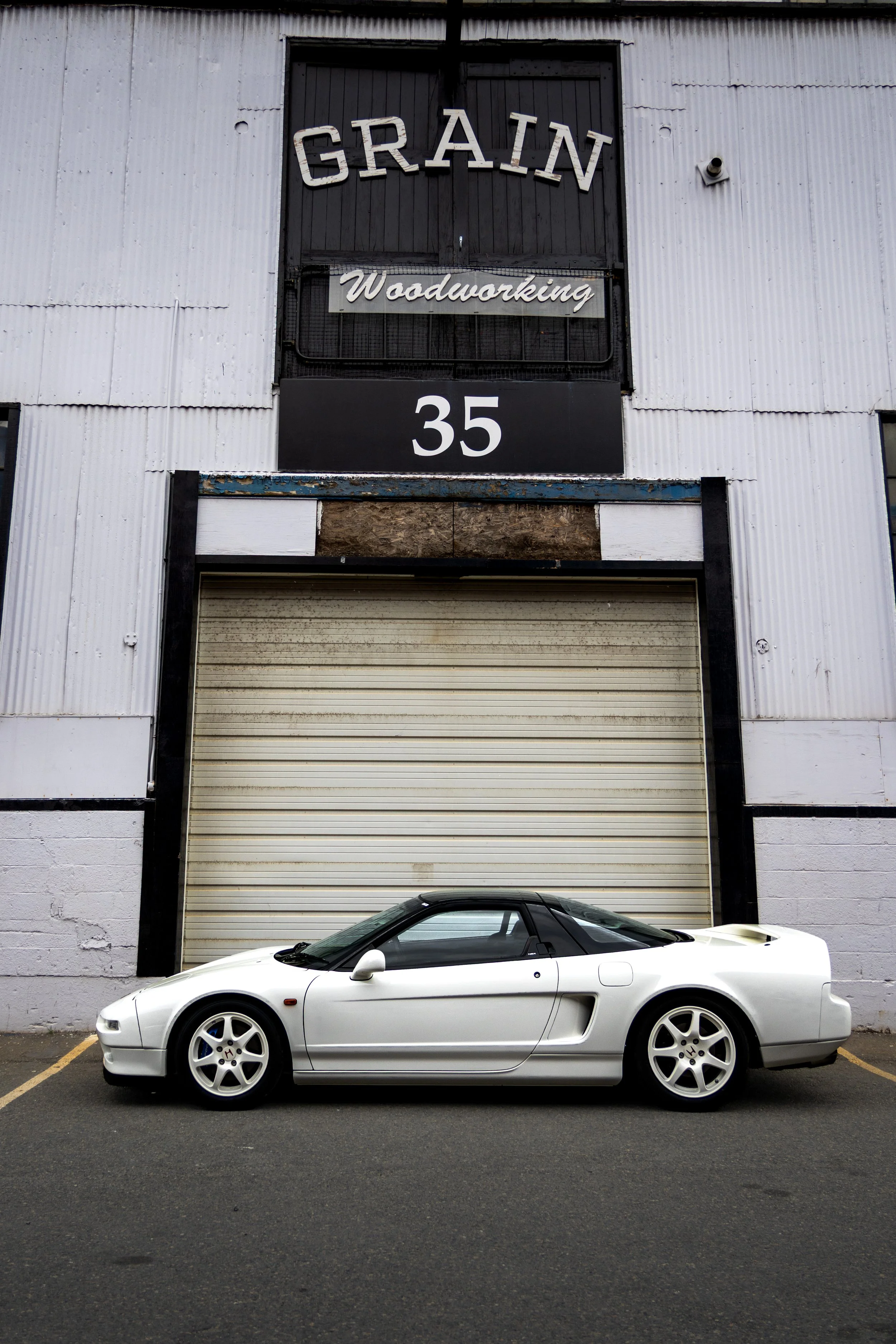 A white sports car parked in front of an industrial building with a closed roll-up door, a sign reading 'GRain' and 'Woodworking,' and the number 35.