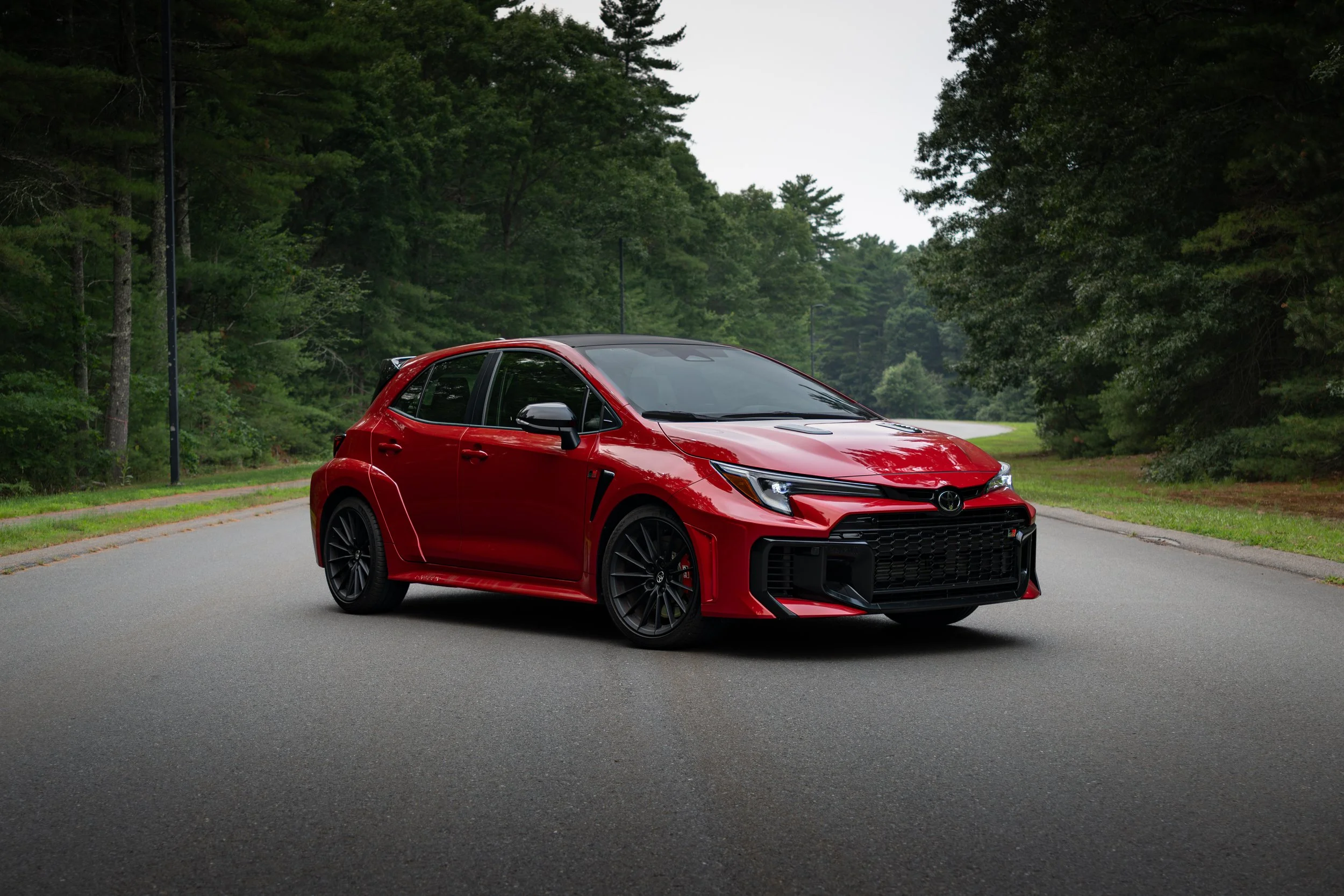 A 2026 Toyota Yaris GR parked on a paved road surrounded by green trees and overcast sky.